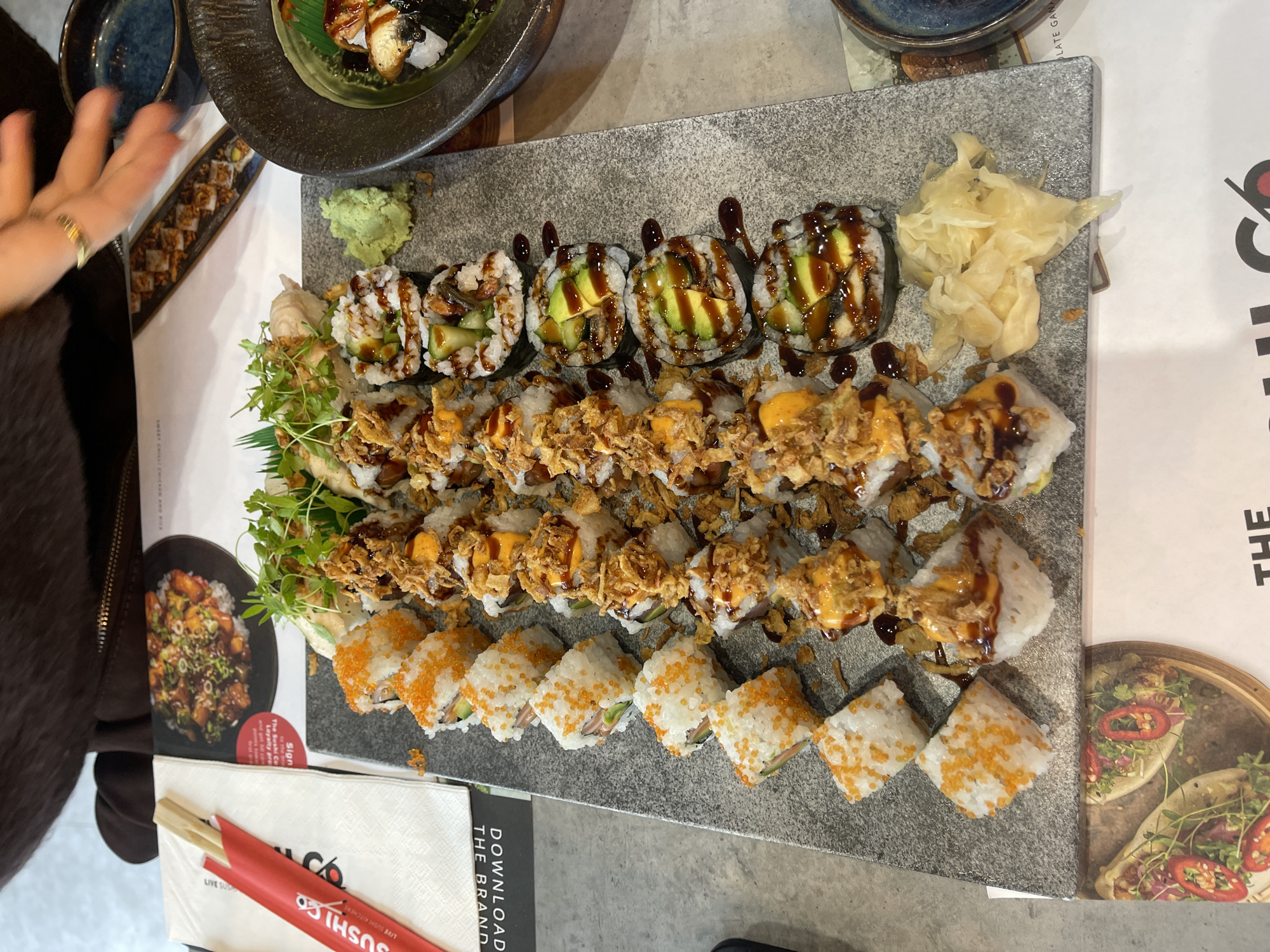 Here is a description of the image:
The image shows a large rectangular grey stone platter filled with different varieties of sushi. The sushi rolls are arranged in rows, with variations featuring toppings like avocado and dark sauce, fried onions and spicy mayo, and orange roe (tobiko). Beside the platter are small containers with accompaniments such as wasabi and pickled ginger. A separate small black dish contains more sushi rolls with what appear to be slices of eel. The platter rests on a table with various menus or promotional materials from "Sushi Co.," a sushi restaurant brand with red chopsticks resting nearby. The scene seems to be inside a restaurant. The exact London location is not identifiable from the image, but the details suggest a contemporary sushi restaurant setting within the city. A hand is visible to the left with gold rings on fingers.