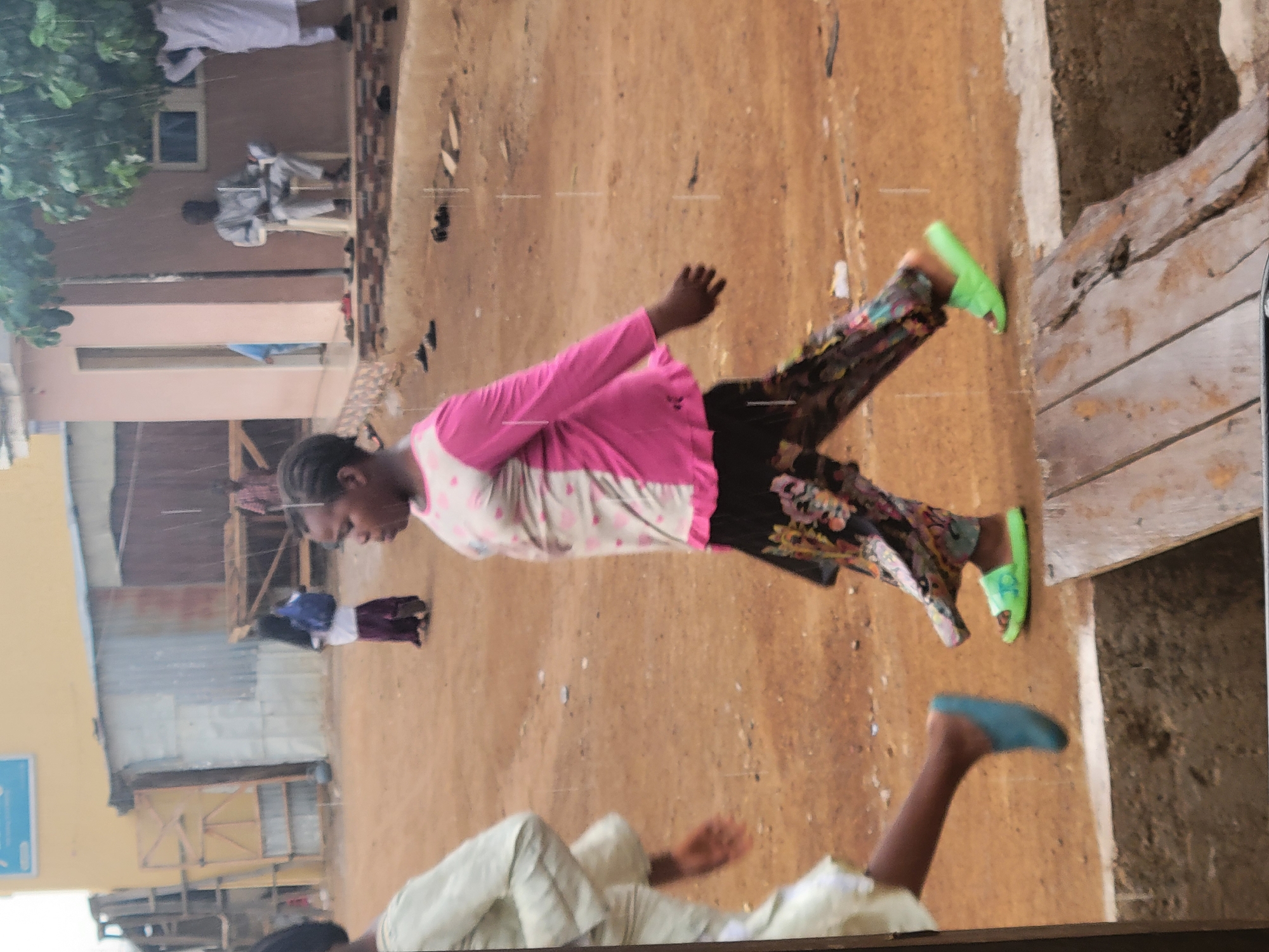 The image captures a scene in Madalla, Nigeria. A woman in a pink and white polka dot top, floral pants, and green sandals walks on a dirt path. Another person wearing a white outfit is partially visible at the bottom. Buildings and other people are visible in the background.