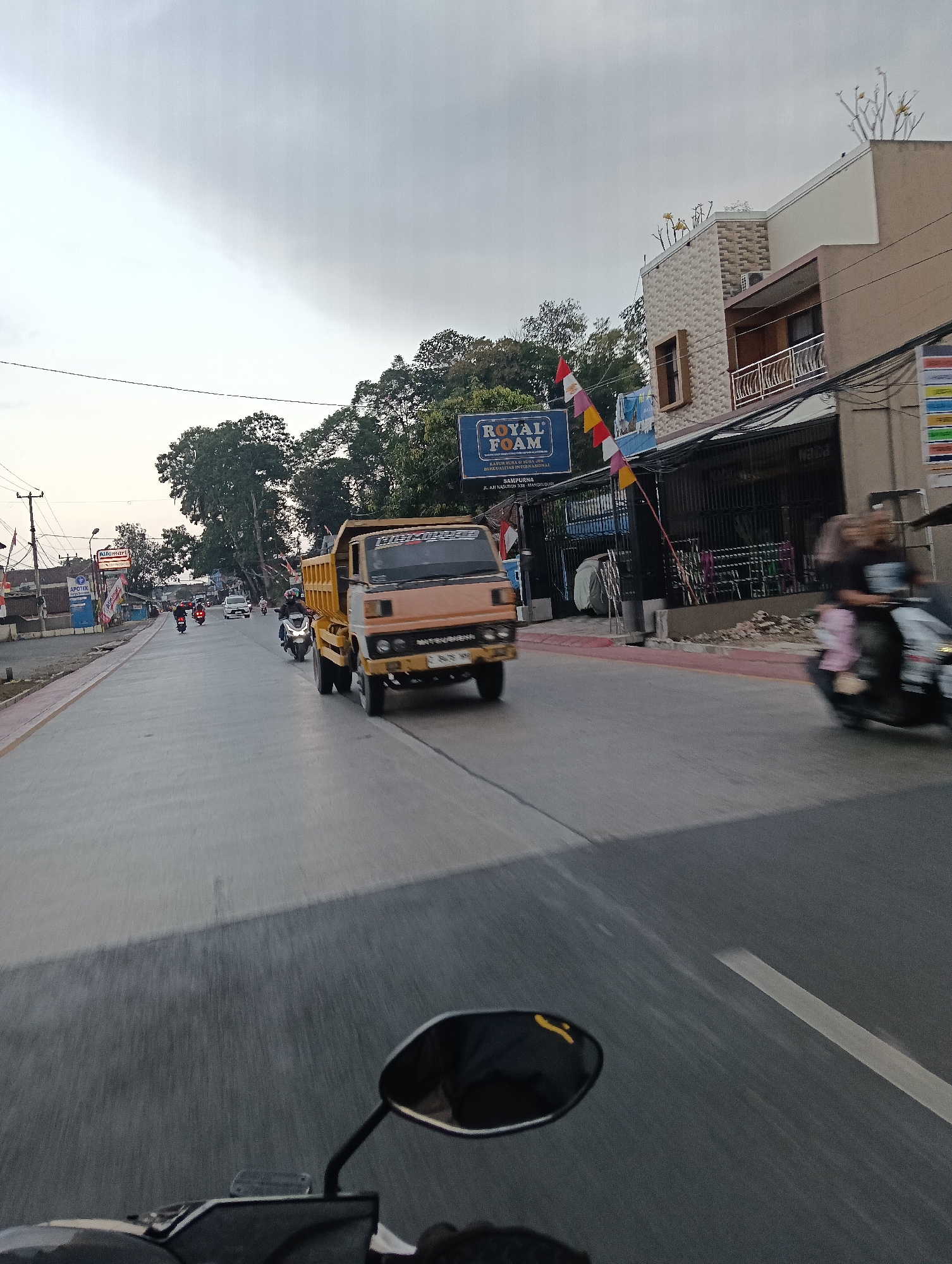The image captures a street scene in Tasikmalaya, Indonesia, likely during the late afternoon or early evening due to the diffused light. In the foreground, the handlebars and a mirror of a motorcycle are visible, suggesting the photo was taken from a moving motorcycle. The road is paved and appears to be a main thoroughfare with multiple lanes.
Dominating the mid-ground is a light orange dump truck traveling forward. To the right of the truck, a motorcycle with two occupants, a woman and a child, is also in motion. Further down the road, several other motorcycles are seen, indicating active local traffic.
Along the right side of the road, there is a building with a modern facade, featuring patterned tiles and balconies. A commercial sign for "ROYAL FOAM" is prominently displayed, along with smaller banners and flags. In the background, trees line the road, and utility poles with wires are visible. The sky is overcast, with a mix of grey and lighter clouds. The overall impression is one of a busy urban environment.