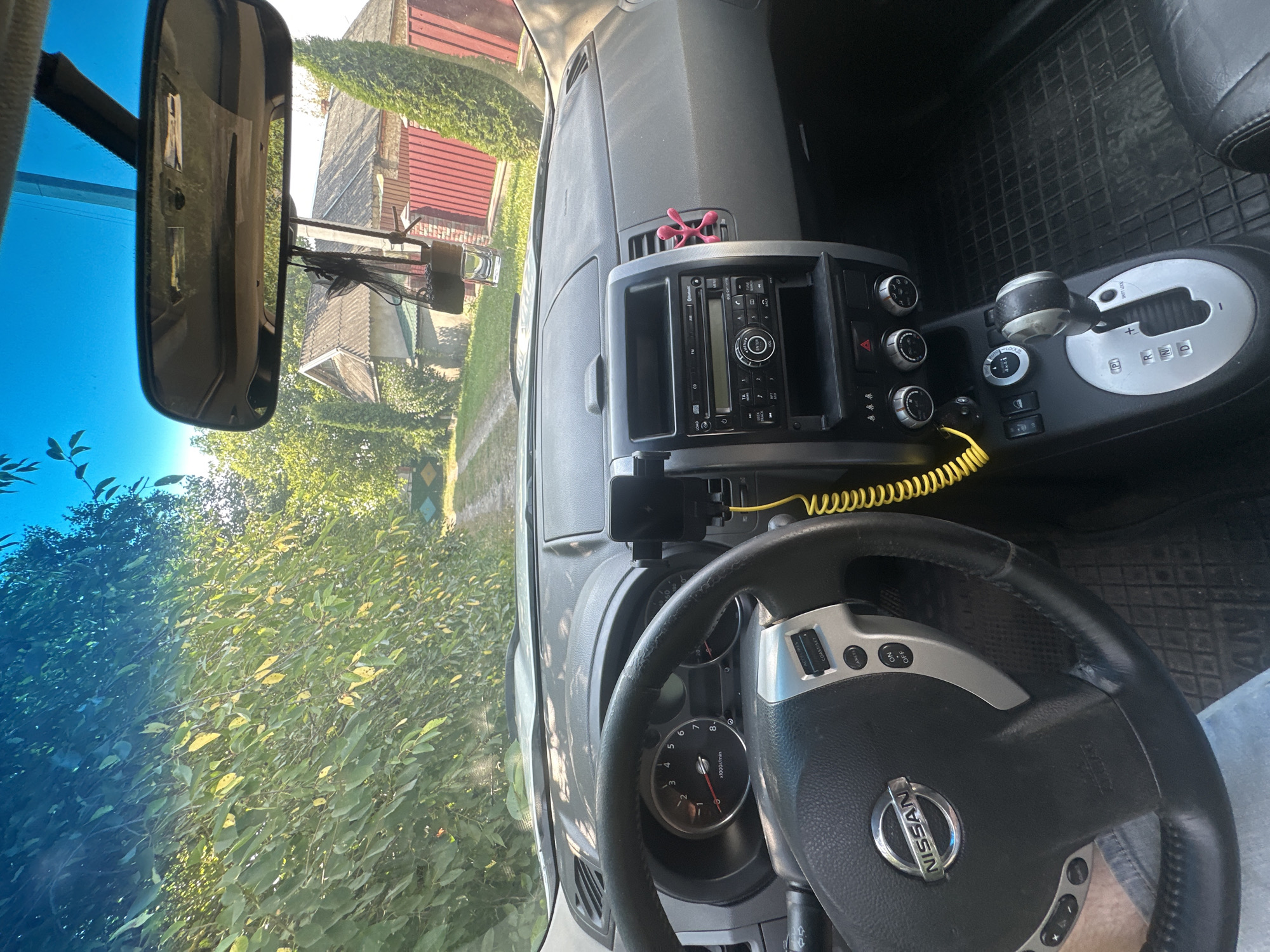The image shows the interior of a Nissan X-Trail parked on a dirt road in Shatava, Ukraine. The sun is shining, creating bright highlights on the dashboard and windshield. Through the windshield, a rural scene is visible, with a dirt road leading past trees and houses with red fences. The interior of the car features a black leather steering wheel with a Nissan logo, a dashboard with various controls including a radio and climate control knobs, and a gear shifter. A yellow coiled cord is visible, likely for charging a device. A small black air freshener hangs from the rearview mirror. A person's leg, wearing denim shorts, is partially visible in the lower left corner.