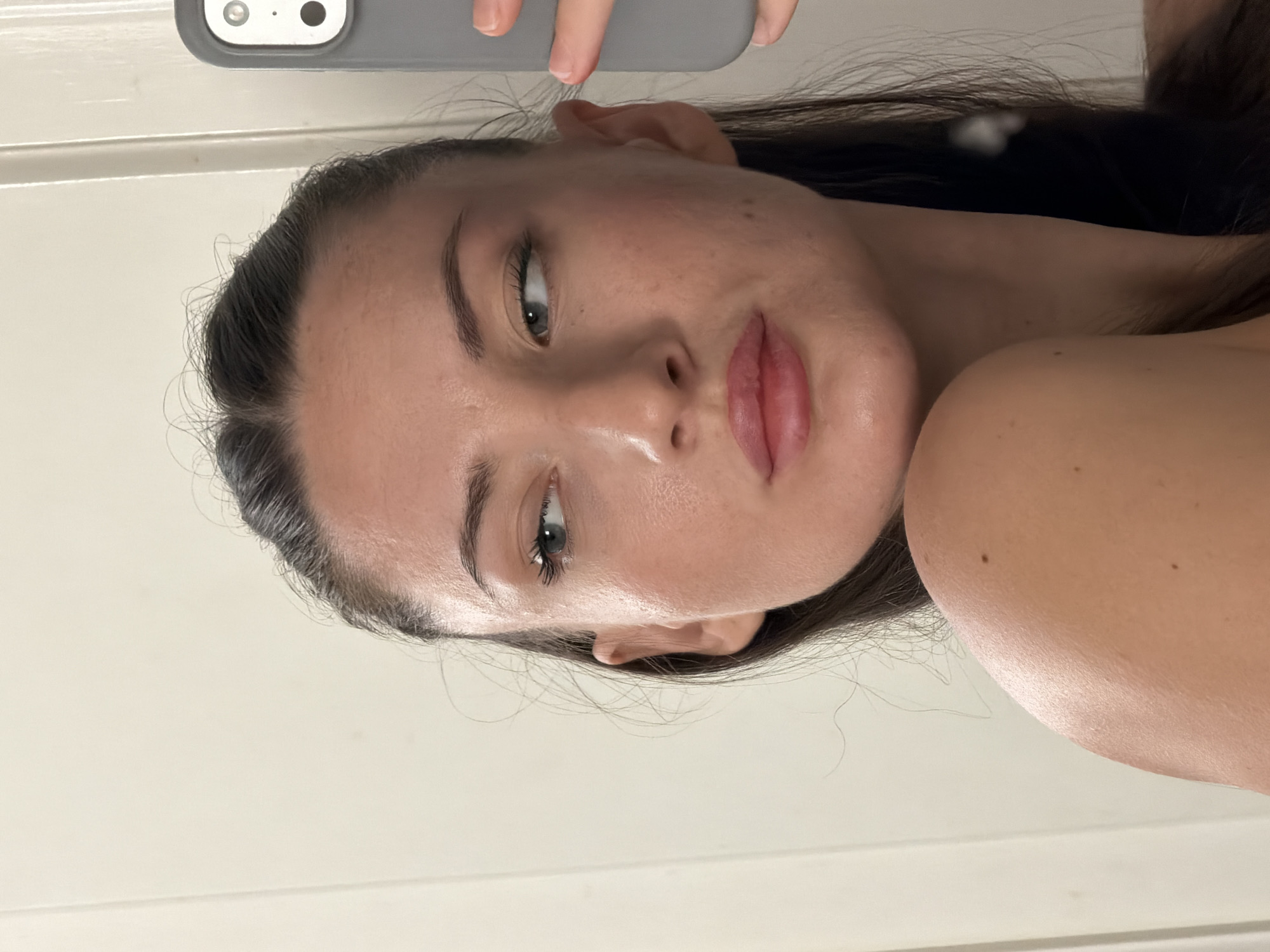 The image shows a close-up, profile view of a young woman's face and upper torso, reflected in a mirror. She has dark hair pulled back, fair skin, light blue eyes, and full lips. She is wearing a light-colored garment that reveals her shoulder and collarbone. She is holding a grey smartphone in her right hand, partially obscuring the mirror's reflection of her face. The background is a plain, light-colored wall with a vertical door frame visible to the left. The lighting appears to be soft and natural. The scene is indoors, likely a bathroom or bedroom, and there are no geographical indicators specific to Sharm El Sheikh, Egypt.