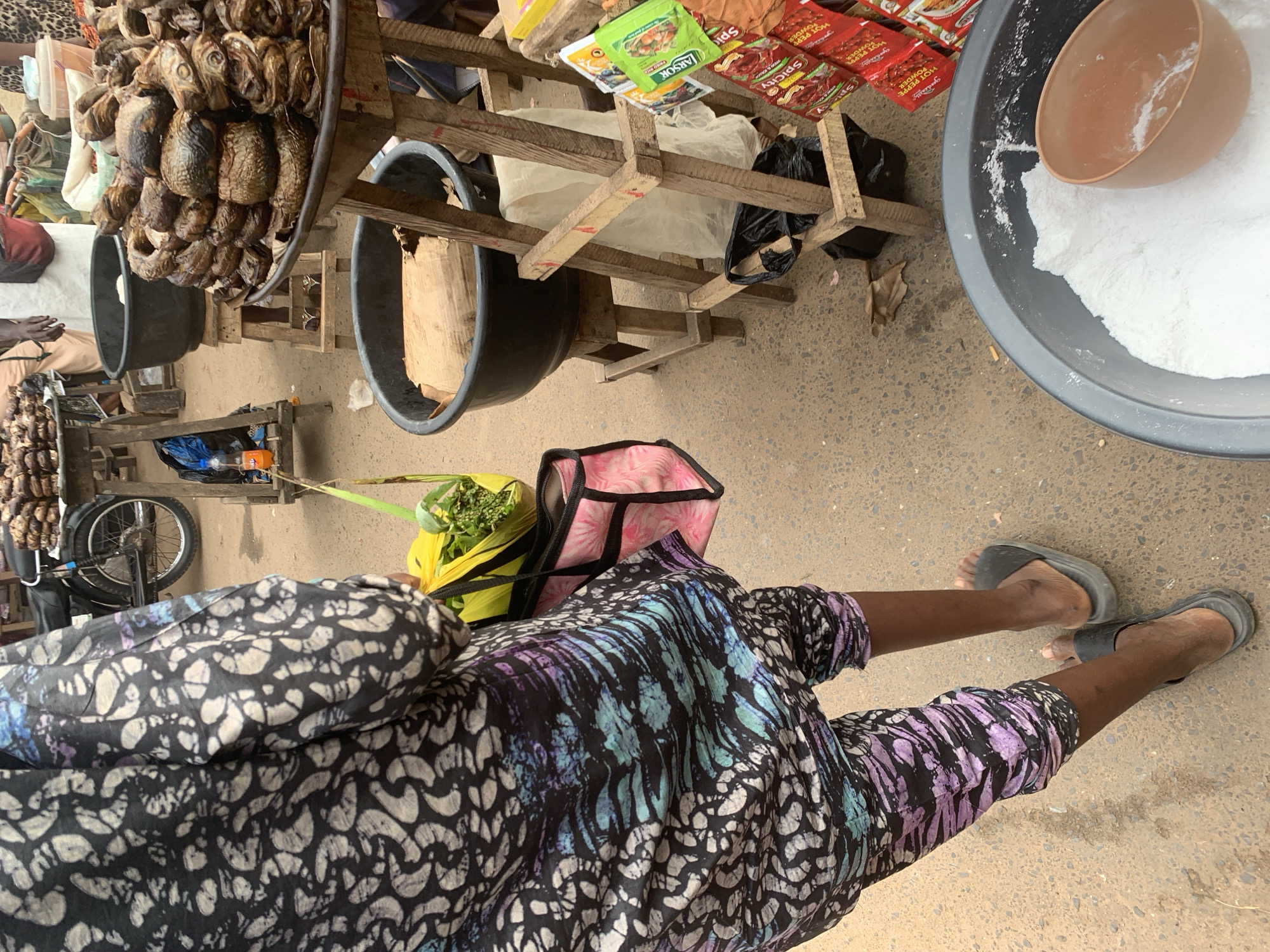 This media file shows a high-angle, eye-level shot of a market scene, likely Shagamu, Nigeria. The focus is on the lower body of a person wearing a vibrantly patterned wrap skirt and dark flip-flops, standing in a marketplace. In front of this person, a yellow bag filled with green produce is visible. Behind them, a pink and black bag rests on the ground.
The background reveals a bustling market environment. Several large dark basins are present, one filled with white powder and a brown scoop, and another empty. Wooden stalls hold dried fish, packaged spices, and other goods. A bicycle is also visible, leaning against a stall. The ground is paved with concrete. The lighting suggests daytime, possibly mid-morning or afternoon, with no indication of specific weather conditions. The scene captures the everyday activity of a marketplace, with goods for sale and people going about their business. No discernible emotions are conveyed, as the faces of individuals are not the primary focus. Some text is visible on the spice packets, including "SPICY" and "HOT PEPPER POWDER."