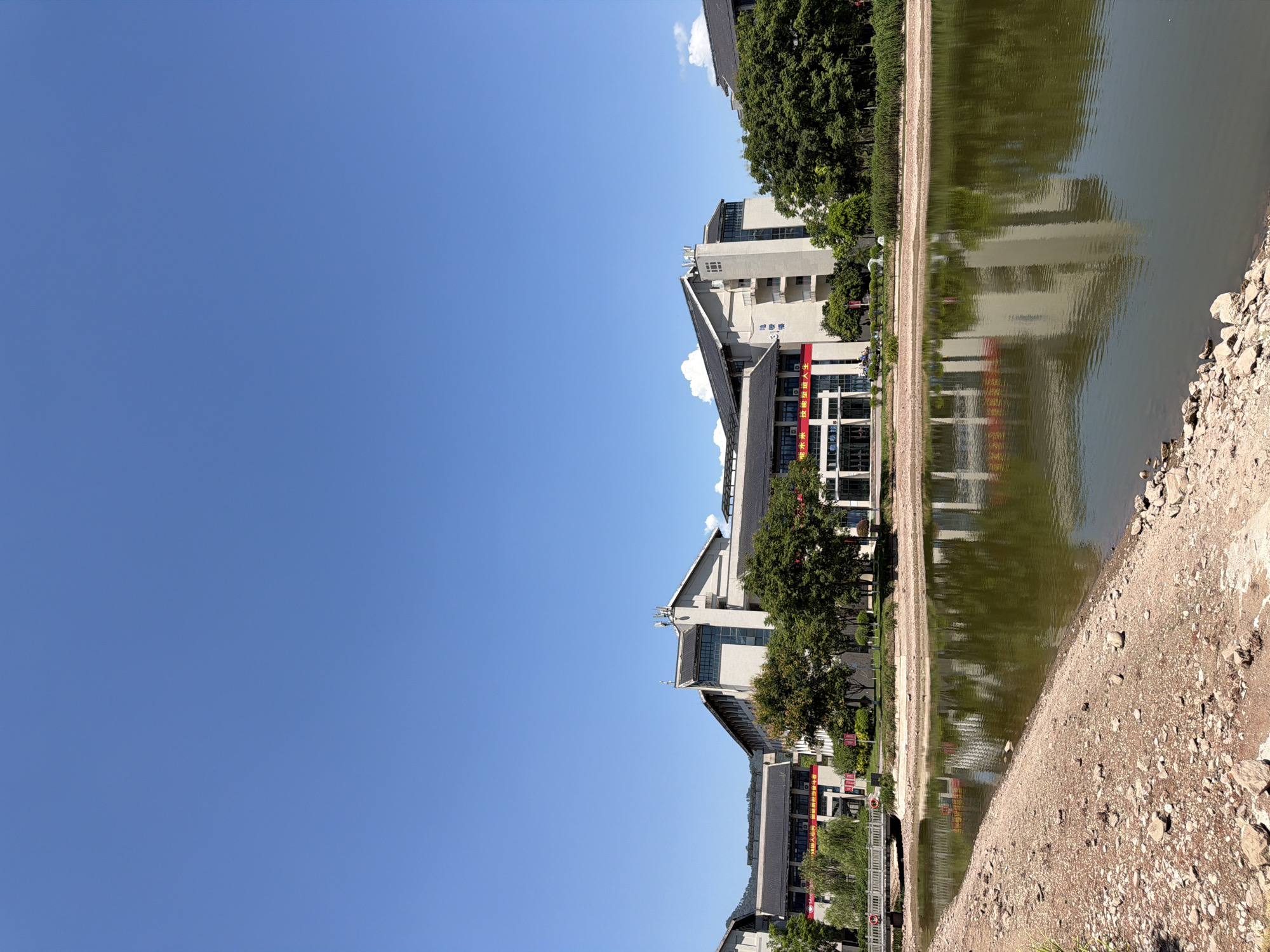 The image depicts a bright, clear day with a vast expanse of blue sky dominating the upper portion. Below, a modern architectural complex with dark tiled roofs is visible, featuring light-colored facades and multiple stories. Red banners with Chinese characters are displayed on some of the buildings. Lush green trees line the front of the buildings, and a body of water, reflecting the architecture and trees, lies in the foreground. The edge of the water features a rocky, barren bank. In the distance, rolling hills are faintly visible against the horizon. A bridge crosses a section of the water near the left side of the frame. The scene appears to be a campus or a public facility in Jiangjin, China.