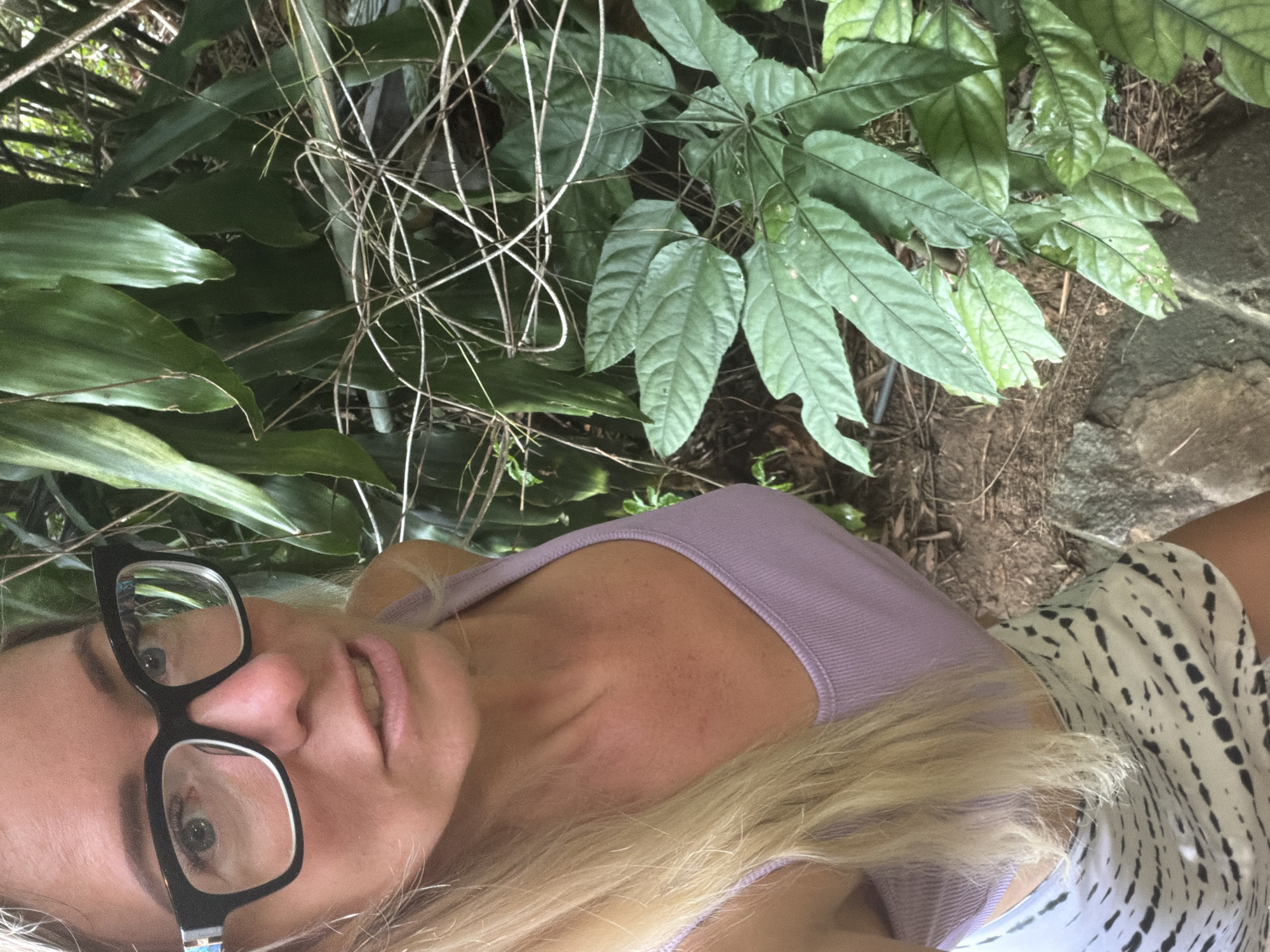In a close-up, low-angle shot, a woman with blonde hair and blue eyes is pictured outdoors, partially obscured by lush green foliage. She is wearing large, black-rimmed glasses and a lilac-colored tank top. Her skirt, visible in the lower portion of the frame, features a black and white tie-dye pattern. The background consists of dense vegetation, suggesting a natural or garden setting, possibly within the city of Xuân Thành, Vietnam. The overall lighting and composition indicate a casual, selfie-style photograph taken amidst the greenery.