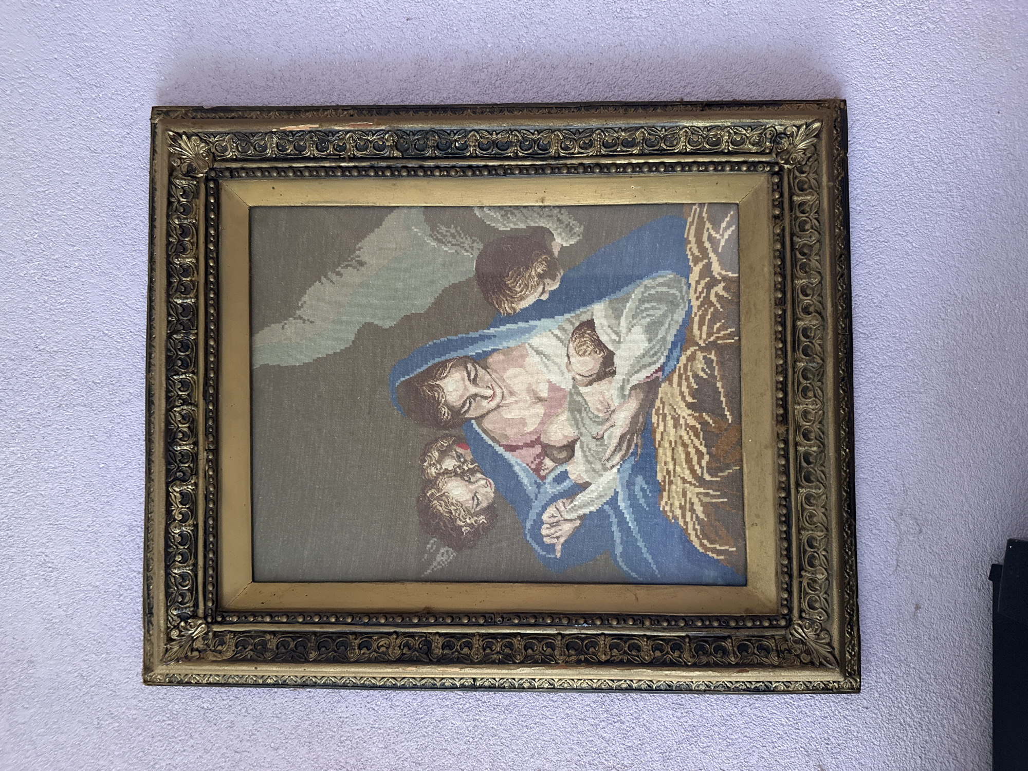 A framed tapestry depicting the Madonna and Child with angels is hanging on a textured, light purple wall. The tapestry shows the Virgin Mary, dressed in a blue mantle and pink tunic, cradling the infant Jesus. Several cherubic figures, possibly angels, are partially visible around them, looking on with adoration. The scene is set against a neutral background, with the figures resting on a bed of straw. The frame is ornate and gold-colored, with detailed carvings. The wall is likely in a domestic setting in the city of Cherven Breg, Bulgaria.