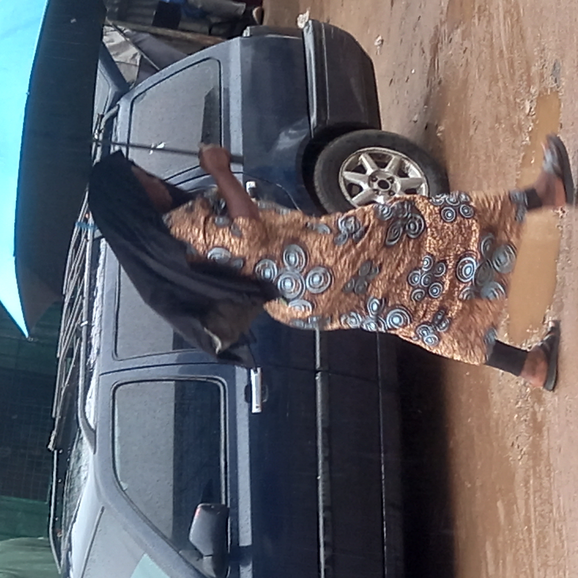 A woman is walking on a wet ground, holding a black umbrella beside a dark blue car on a street in Dadi Riba, Nigeria. The woman wears a brown and blue patterned dress over black leggings and black sandals. The car has a roof rack.