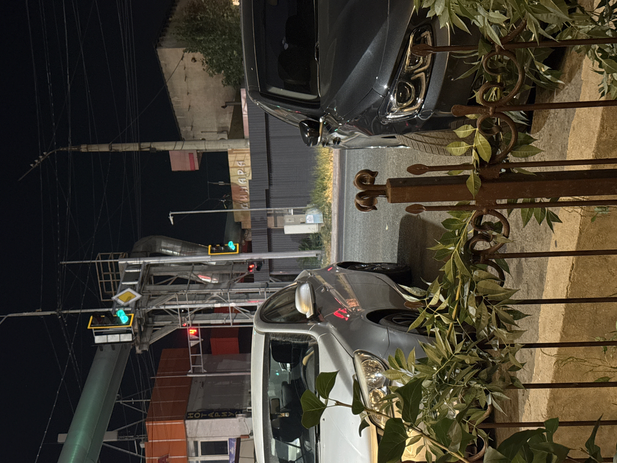 The image depicts a nighttime street scene in Shymkent, Kazakhstan, likely near an intersection. Two cars are parked side-by-side on the right side of the frame. The car on the left is silver, and the car on the right is dark gray. In the background, traffic lights are visible, with a green light illuminated on one. There are also overhead wires and a large metallic structure, possibly part of an elevated walkway or industrial equipment. A building with an orange facade is on the left, and a lighter-colored building with a textured wall is on the right. A metal fence with ornate scrollwork and green foliage in front of it occupies the foreground, partially obscuring the cars. The overall impression is of an urban environment at night, with traffic infrastructure and parked vehicles.