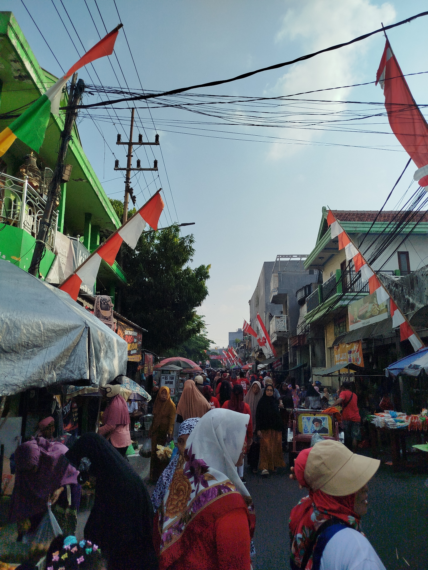 The image depicts a street scene in Surabaya, Indonesia, likely during a public event or festival given the decorations. Red and white flags, common for Indonesian national celebrations, are prominently displayed along the street, hung from buildings and utility poles. The street is lined with shops and stalls, some covered with tarps. A crowd of people, many wearing headscarves, are walking along the street, suggesting it is a busy thoroughfare. The architecture of the buildings suggests a mix of residential and commercial structures, typical of urban areas in Indonesia. The sky is clear with some scattered clouds. The overall impression is of a lively, populated urban environment.