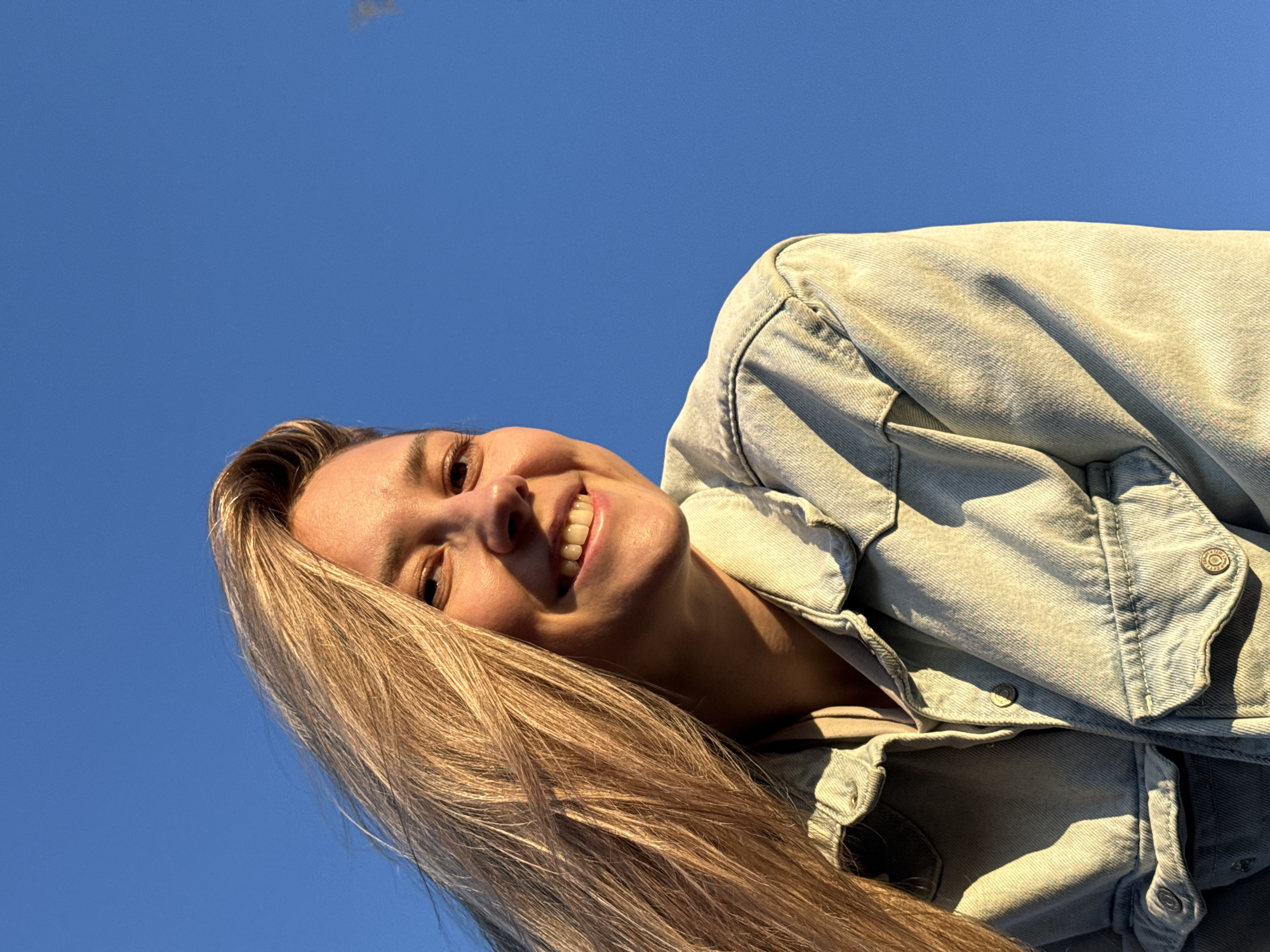 A young woman is smiling broadly while looking down at the camera. She has long, blonde hair. She is wearing a light-colored denim jacket. The background is a clear, blue sky. There is no information in the image to confirm the location is Chaikovsky, Russia.