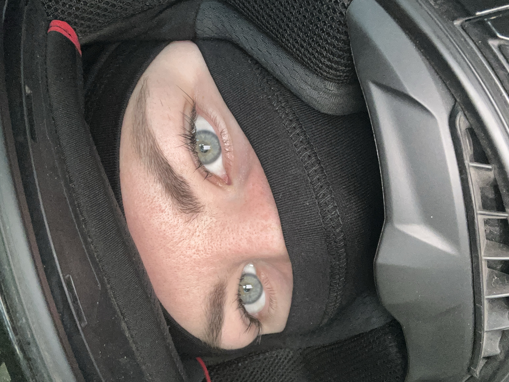 The image presents a close-up, rotated view of a person's face, focusing on their eyes and upper cheeks, peering out from within a black helmet interior. The person wears a snug-fitting black balaclava or head covering that conceals the hair and lower face, allowing only the eyes and a segment of the forehead and upper cheeks to be seen.
The eyes are a prominent feature, displaying a light blue-grey hue with visible, long, dark eyelashes and dark, well-defined eyebrows. The skin visible is fair, with a subtle texture of pores or light freckles on the cheeks. The expression appears neutral, with the gaze directed forward.
The surrounding elements are components of a helmet interior, featuring various black textures. There is soft, padded black fabric cushioning directly around the face, transitioning to a more textured, mesh-like material in other areas. Hard, matte black plastic elements, possibly part of the helmet's ventilation or structural design, are visible on the right side. A small red detail, possibly a strap or tag, is discernible on the far left side of the frame, contrasting with the dominant black tones. There is no visible text in the image. The setting suggests the person is prepared for an activity requiring head protection, but the specific location, time of day, or weather are not identifiable.