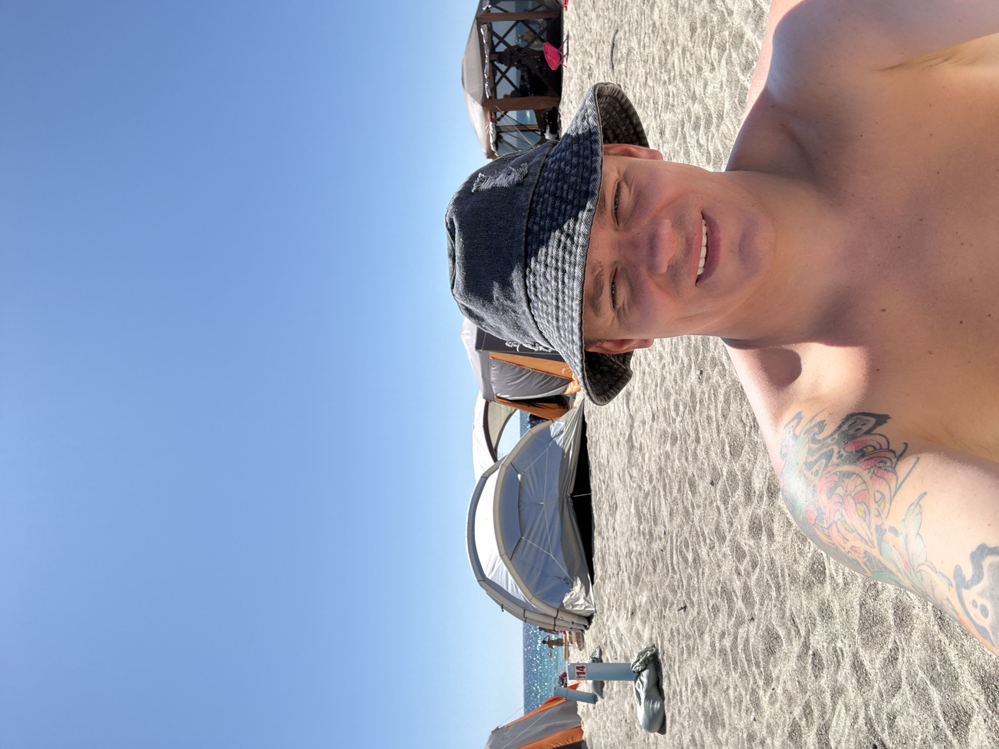 A man wearing a bucket hat and no shirt is taking a selfie on a sandy beach. He has tattoos on his left shoulder and arm. In the background, there are several tents set up, with the ocean and a clear blue sky visible beyond. The beach is populated with a few other people in the distance. The scene appears to be a campsite on a sunny day. The location is not identifiable as Nakhodka, Russia based on the visual information in the image.