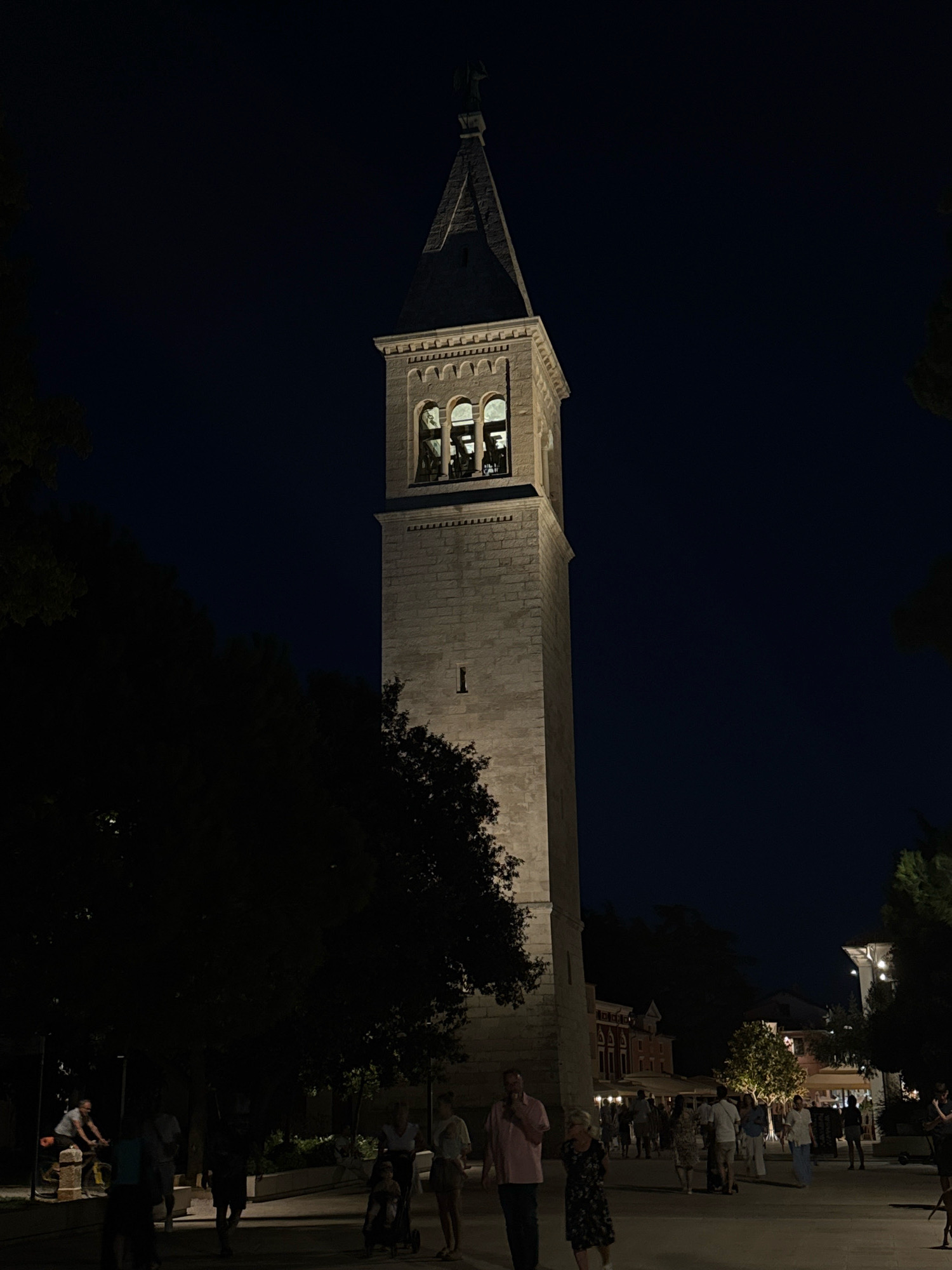 The image depicts a nighttime scene in the city of Novigrad, Croatia, featuring a prominent stone bell tower as its central focus. The tower, illuminated by artificial lights, displays arched windows at its belfry and a pointed roof, topped with a statue. Surrounding the tower, a square or plaza is visible, bustling with pedestrians engaged in various activities. People are seen walking, some are with children in strollers, and a cyclist is present on the left side of the image. The background reveals buildings with illuminated windows, suggesting a lively urban environment. Trees are scattered throughout the scene, some casting dark shadows. The overall atmosphere is one of a pleasant evening outing, with the bell tower serving as a landmark.