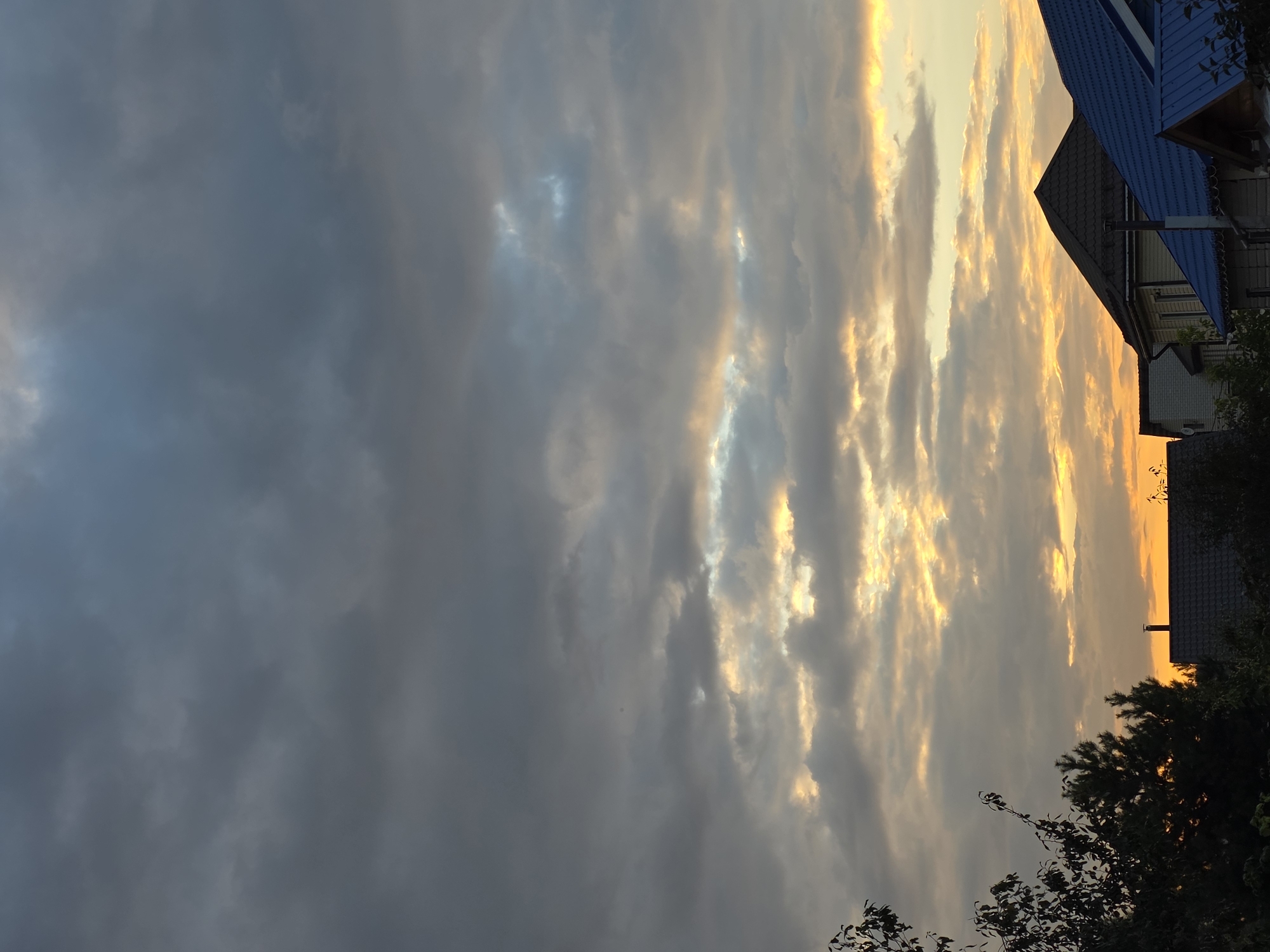 The image depicts a twilight scene over a residential area in Rechytsy, Russia. The sky is filled with dramatic, layered clouds, ranging from dark grey at the top to lighter, more illuminated clouds closer to the horizon. The setting sun casts a warm, golden glow through breaks in the clouds, creating streaks of orange and yellow light.
Below the sky, the rooftops of several houses are visible. These houses have varied roof designs, including dark grey shingles and bright blue metal roofing. Chimneys are present on some of the houses. The foreground is partially obscured by the dark silhouettes of trees and foliage, suggesting a natural environment adjacent to the residential area. The overall atmosphere is serene and captures the transition from day to night. No people are visible in the image.