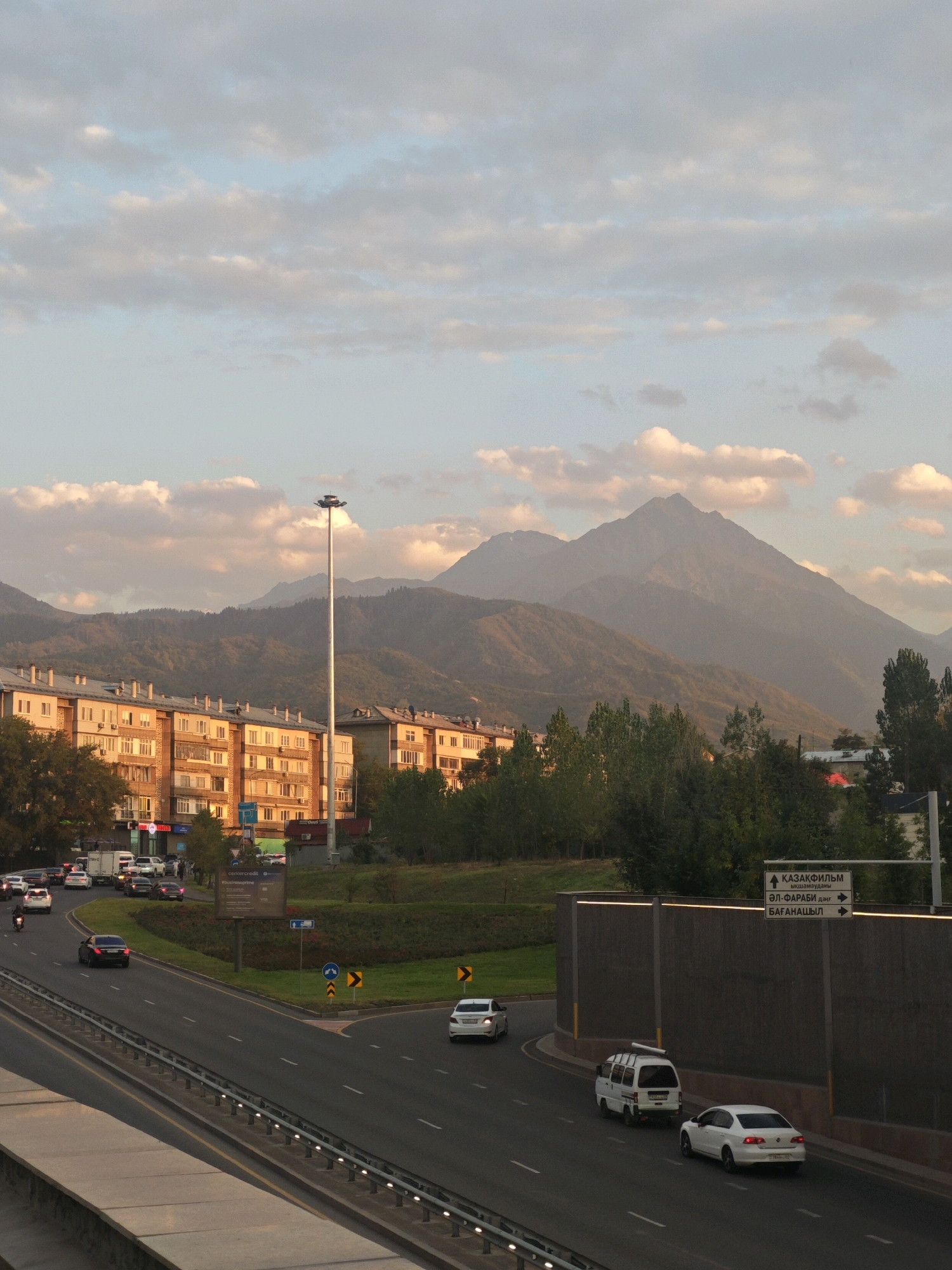 The image depicts a street scene in Almaty, Kazakhstan, with a view of the Tian Shan mountains in the background. The foreground shows a multi-lane road with several vehicles, including cars, a van, and a motorcycle. A white sedan is parked on the right side of the road. In the middle ground, there are apartment buildings and trees. A tall streetlamp stands near the apartment buildings. The sky is partly cloudy, with soft pink and orange hues from the setting sun illuminating the clouds. A signpost with Cyrillic lettering indicates directions to "ҚАЗАҚФИЛЬМ" and other locations. The overall atmosphere is calm and serene, suggesting the late afternoon or early evening.