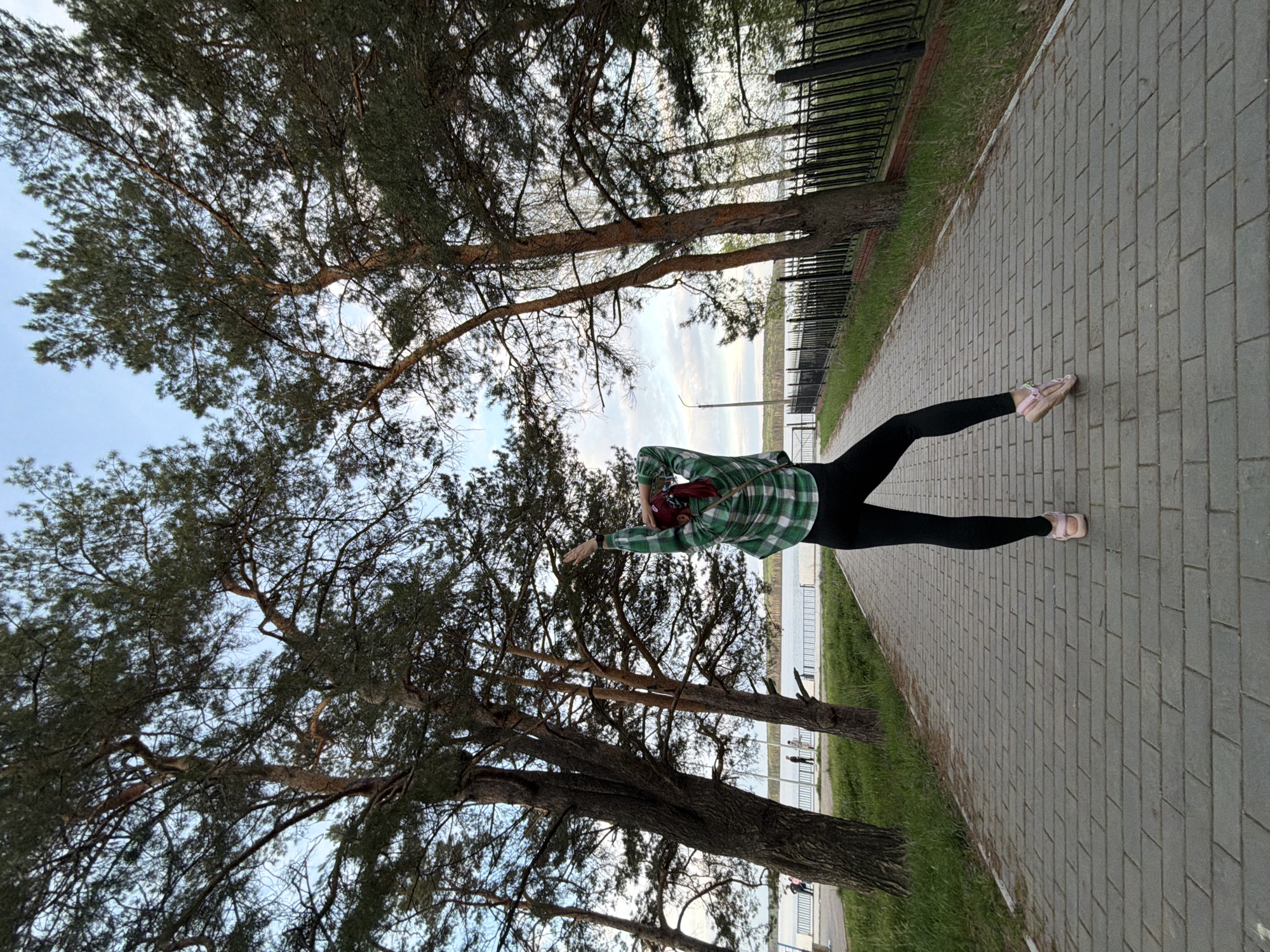 A woman in black leggings and a green plaid shirt stands on a brick walkway in a park-like setting, possibly in Chaikovsky, Russia. She is positioned between tall pine trees with a view towards a body of water, possibly a lake or river, in the distance. A metal fence runs along the edge of the walkway, separating it from a grassy area. The woman is in a dynamic pose, reaching out with her arms, possibly engaging in a stretching or dancing motion.