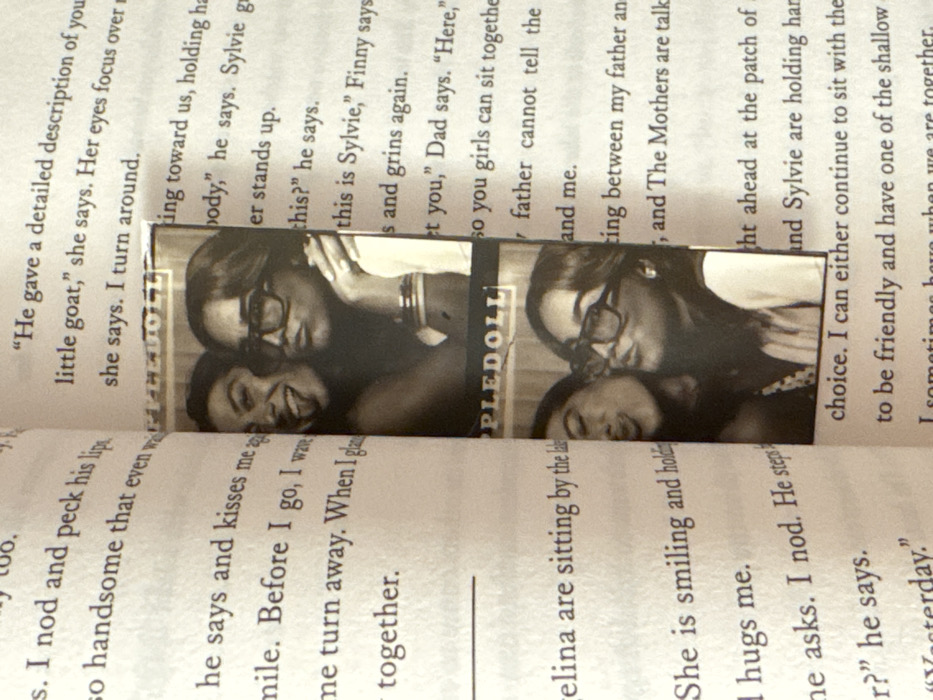 An open book, identified as a novel with cream-colored pages and black serif text, is depicted with a two-panel photo strip serving as a bookmark. The photo strip is positioned vertically between the open pages. The pages display partial sentences; for instance, the left page includes "I nod and peck his lips" and "Angelina are sitting by the la," while the right page reads "He gave a detailed description of yo" and "this is Sylvie,' Finny says."
The photo strip, in a black and white or sepia tone, shows two distinct head-and-shoulder photographs of two young adult females. The top of the strip has partially visible text "PPLEDOLI."
In the upper photo panel, two females are posing together. The individual on the left has dark hair, a wide smile, and her tongue slightly extended. The individual on the right has dark hair, wears large, square-framed glasses, and has her lips pursed in a "duck face" expression. Both are looking towards the camera, positioned closely side-by-side.
The lower photo panel features the same two females. The individual on the left is turned towards the right, puckering her lips as if to kiss the cheek of the other person. The individual on the right, wearing the same glasses, also has her lips pursed in a "duck face" pose, looking towards the camera. Both individuals are wearing bangles on their right wrists. The background in both photo panels appears uniform, consistent with a photo booth setting.
The scene is located in the city of Los Angeles, United States of America.