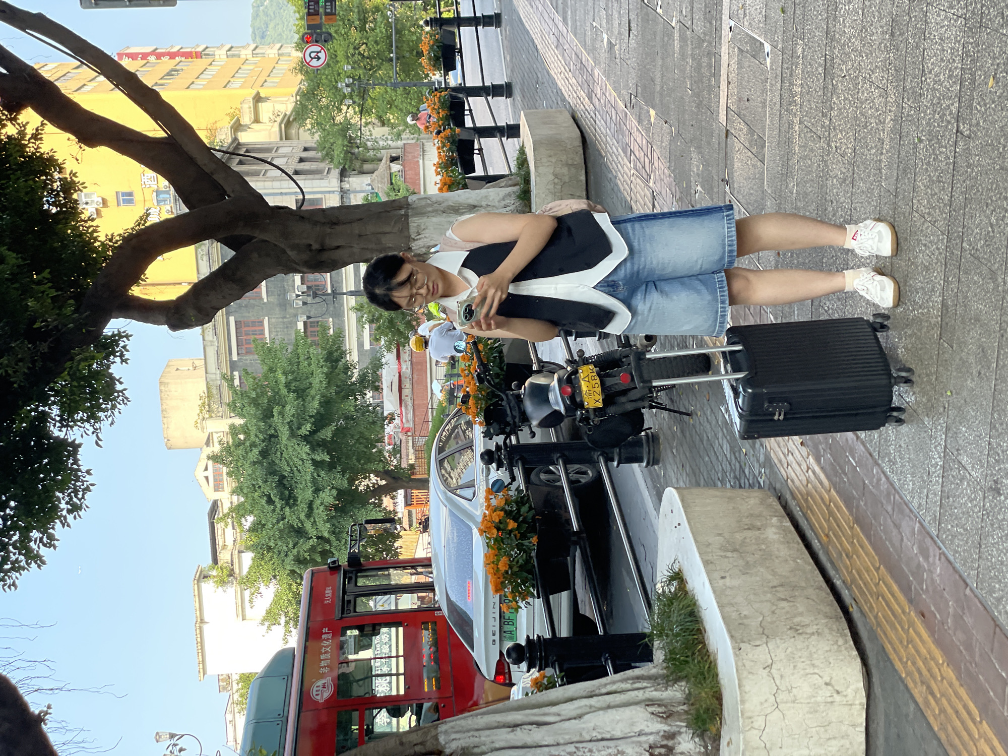 A young woman with glasses stands on a sidewalk in Yuzhong District, China, looking at her phone. She is wearing a white sleeveless top, a dark vest, denim shorts, white sneakers, and ankle socks. Next to her is a black rolling suitcase. In the background, a red bus, a white car, and a motorcycle are visible. The street is lined with trees and planters filled with orange flowers. Buildings, including a yellow high-rise, are in the distance. The sidewalk has tactile paving for the visually impaired.