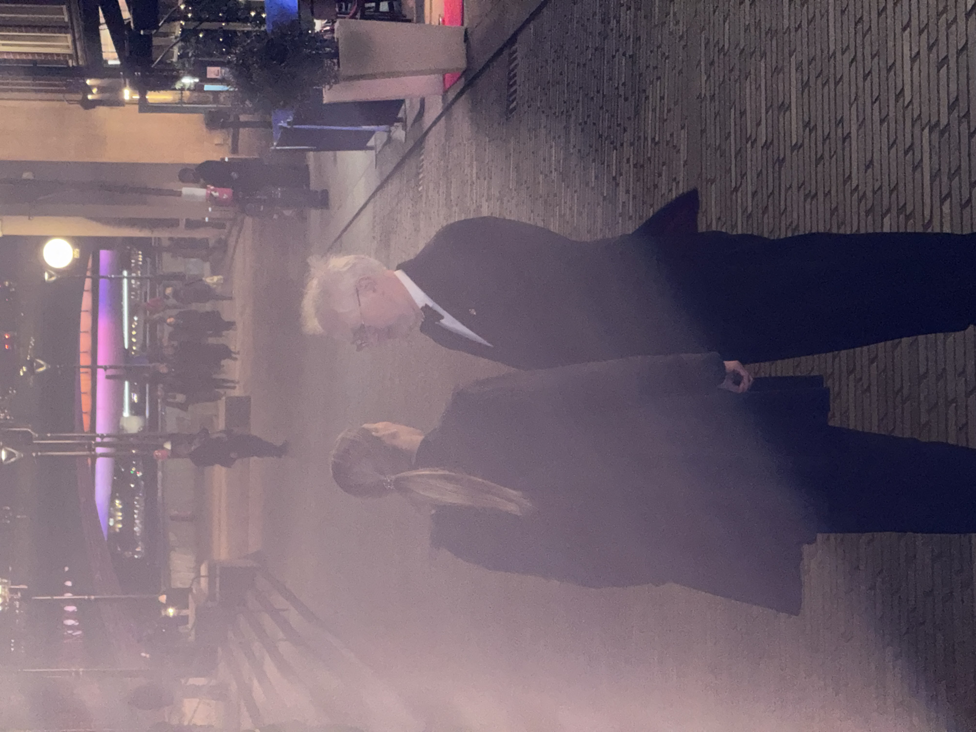 An evening image captures a section of the South Bank promenade in London, United Kingdom, overlooking the River Thames. The Millennium Bridge, illuminated with prominent purple and orange lights, is visible in the background, connecting to the opposite bank. In the immediate foreground, two individuals are positioned on a paved walkway. A person with light-colored hair, tied in a ponytail, stands with their back mostly to the camera, wearing a dark overcoat and trousers. Facing this individual is a male with white hair, glasses, dressed in a dark tuxedo with a white shirt and black bow tie, a small pin visible on his lapel. Both appear to be engaged in conversation. A slight atmospheric haze or fog diffuses the light throughout the scene. To the left, a set of stone steps leads down to the promenade, with blurred figures ascending and descending. On the right, a building features outdoor dining or lounge areas with dark awnings, potted greenery, and a red carpet extending from an entrance, flanked by large, light-colored planters. Additional blurry pedestrians and streetlights are scattered in the midground and background.