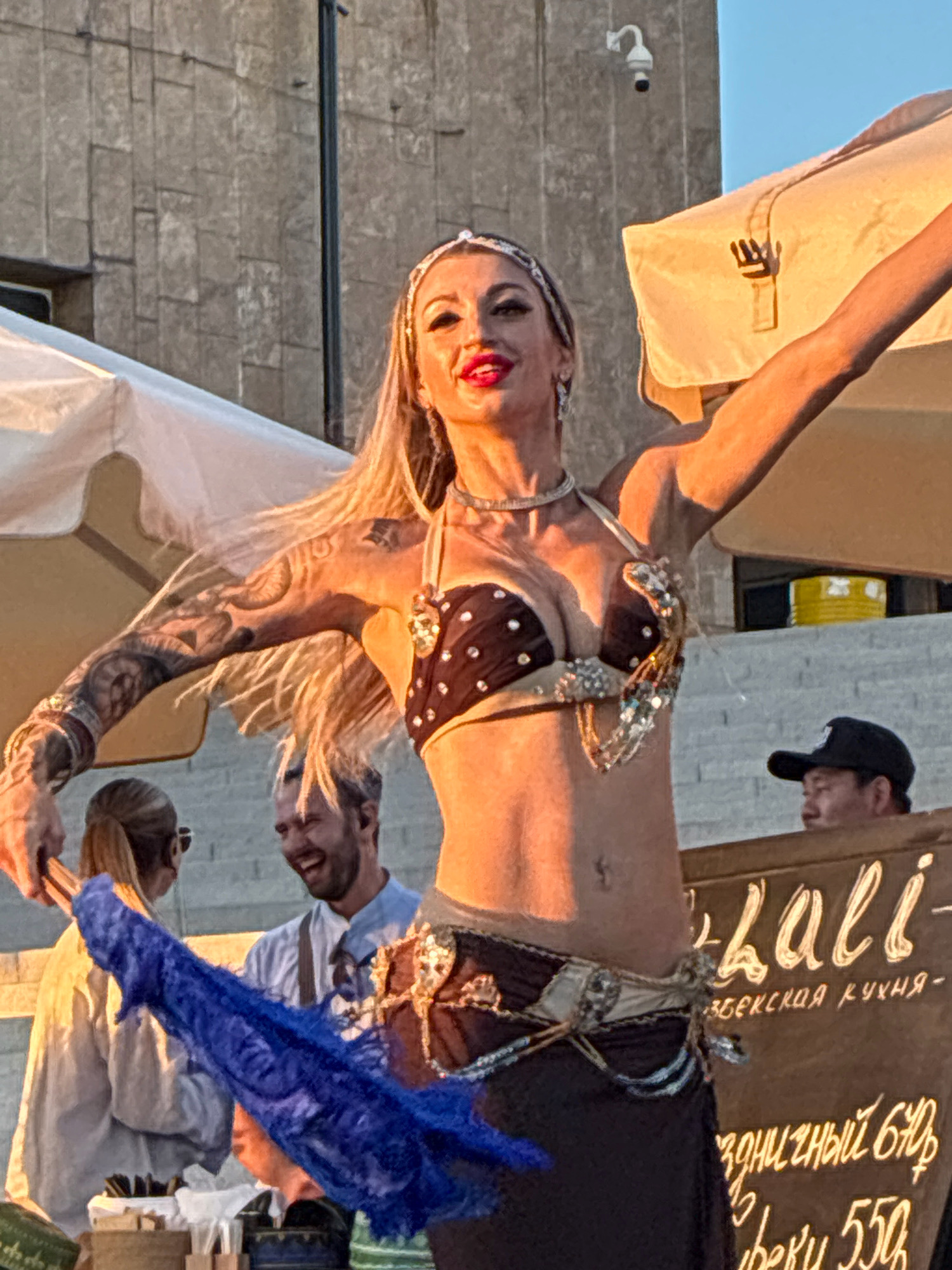 In Sochi, Russia, a female belly dancer performs in front of a stone wall and a tent, bathed in warm sunlight. She wears a dark, sequined bikini top adorned with silver and gold accents, a jeweled belt, and a dark skirt. Her blonde hair flows, and she has bold makeup. She holds a blue lace fan in her left hand and has tattoos on her right arm and torso. In the background, a man with graying hair laughs, and a person with blonde hair tied in a ponytail stands to the left. A man in a black baseball cap observes from behind a wooden counter on the right, near a sign with Cyrillic lettering and a depiction of the raised fist symbol.