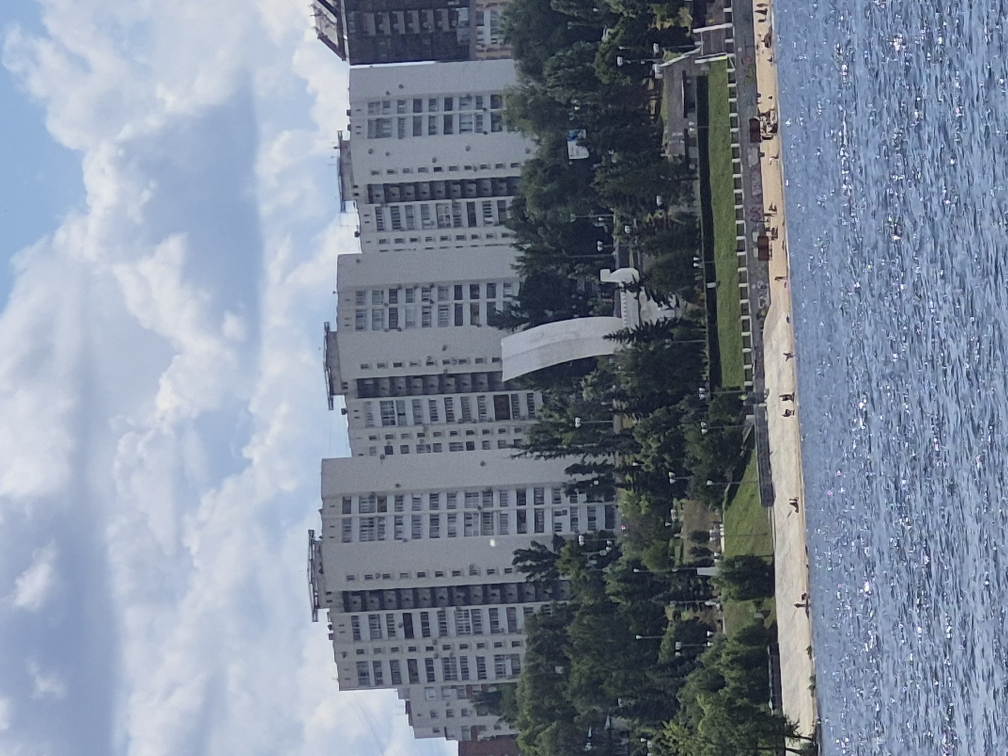 This is a medium-distance, slightly elevated shot of a riverbank scene in Samara, Russia. In the foreground, the choppy, sun-dappled surface of the Volga River dominates the lower half of the image. Beyond the river, a concrete embankment and a sandy beach are visible. A few scattered people are present on the beach, some appearing to be sitting or lying down.
Behind the beach, a sloping green hill is covered with numerous trees of varying heights, interspersed with lampposts. Further up the hill, several tall, light-colored apartment buildings are clustered together, forming a distinctive skyline. These buildings have multiple floors and many windows. To the left of the apartment buildings, a large, white, curved monument stands prominently on the hillside. To the far right, a darker, taller building rises, partially obscuring the sky. The sky itself is partly cloudy, with patches of blue sky visible between the white cumulus clouds.