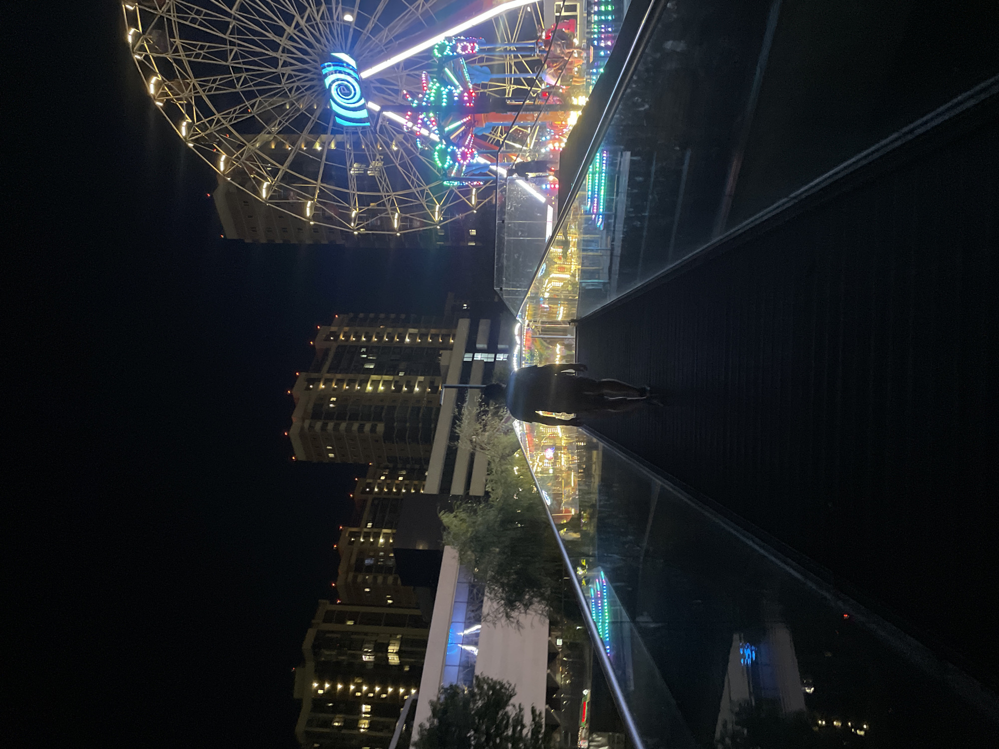 A lone figure walks up a pedestrian walkway at night. The walkway has a glass railing illuminated with colorful lights, and it leads towards a brightly lit amusement park with a Ferris wheel. In the background, several tall buildings with illuminated windows suggest an urban setting. The scene appears to be located at a waterfront, with reflections visible in the water below the walkway. The overall atmosphere is one of urban nightlife and entertainment.