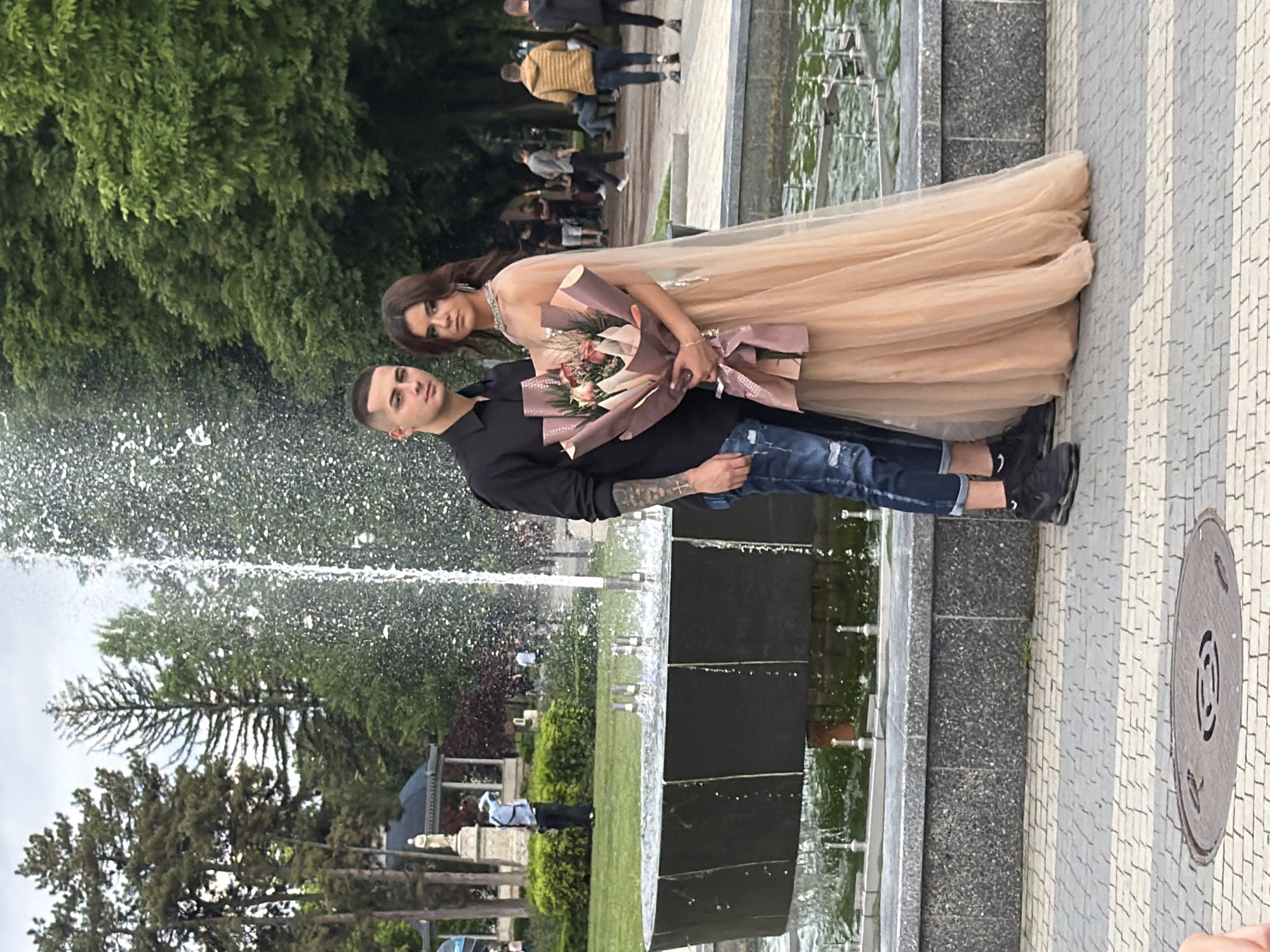 A photograph captures a couple posing in front of a fountain. The woman is wearing a long, tan-colored gown and holding a bouquet of flowers wrapped in brown paper with a large brown bow. The man is wearing a black shirt and blue jeans. They are both looking towards the camera.
The fountain is situated in a park or public space, with green grass, trees, and other vegetation visible in the background. A number of people can be seen walking around in the background. The location is in Pleven, Bulgaria.