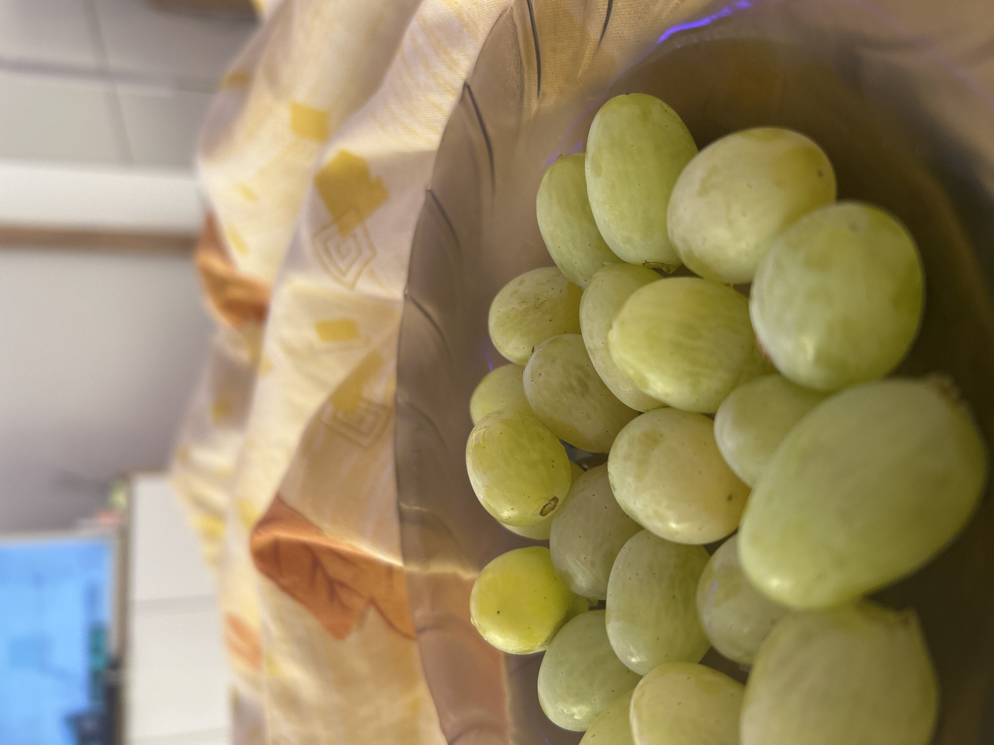 A close-up, eye-level shot displays a clear glass bowl filled with light green grapes. The grapes are clustered together, with some in sharp focus and others softly blurred. The bowl itself has subtle iridescent qualities, catching the light. Behind the bowl, a yellow and orange patterned fabric is draped, appearing to be a bedspread or tablecloth, with soft folds and wrinkles. In the background, out of focus, is a white surface, possibly a dresser or cabinet, topped with a blurred object that suggests a television screen or monitor. The overall lighting is soft and natural, creating a relaxed and intimate atmosphere. The location is identified as Malo Konare, Bulgaria.