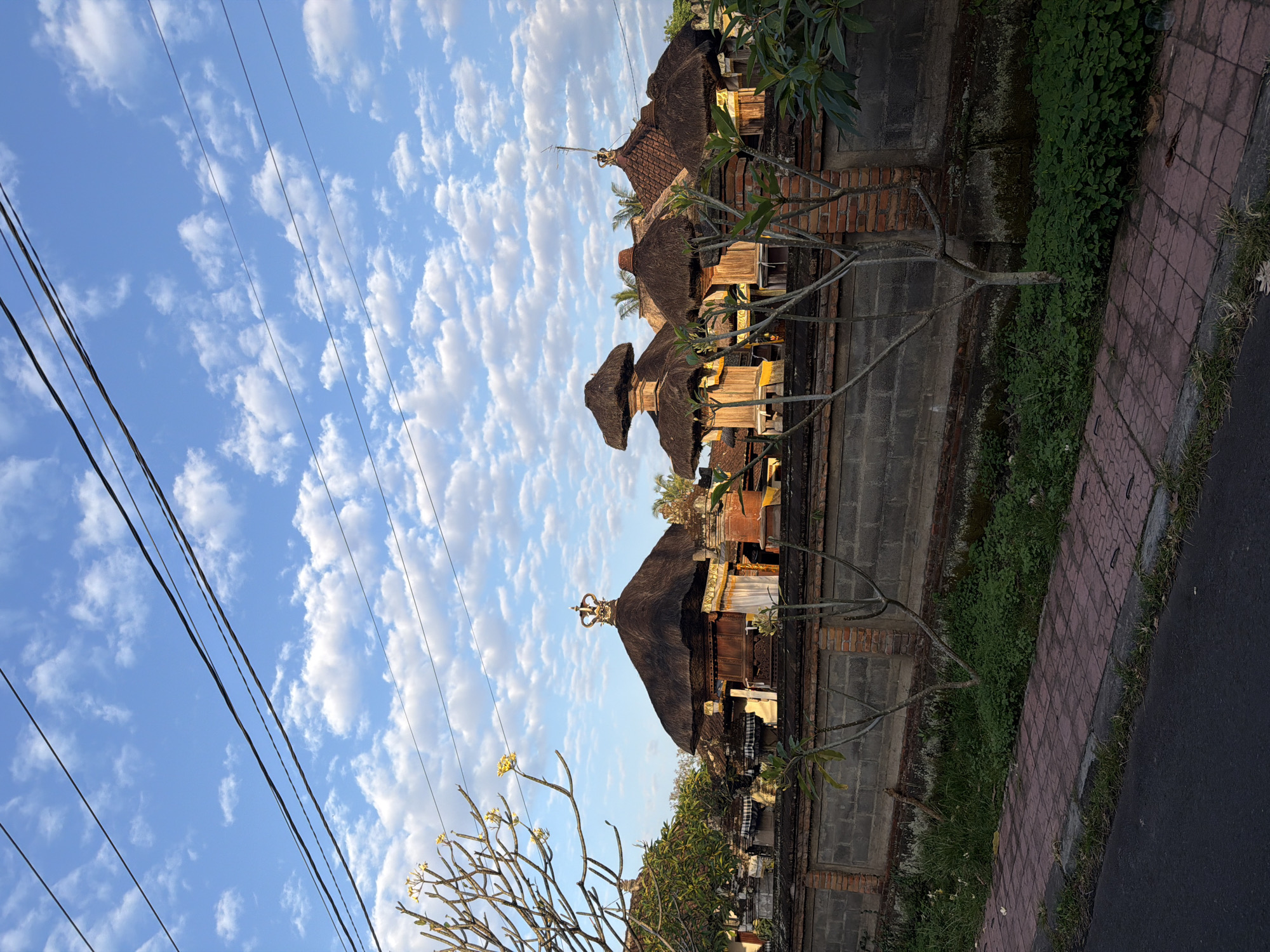 The image depicts a view of several traditional Balinese structures with thatched roofs, likely part of a temple complex or a residential compound, located in the Ubud District, Indonesia. The buildings are characterized by their tiered roofing and ornate crowns atop some of the structures. A brick and concrete wall separates the buildings from the foreground, which features a paved sidewalk with red tiles and a dark asphalt road. Green vegetation, including small trees and shrubs, grows along the wall and sidewalk. The sky above is a vibrant blue, dotted with scattered white clouds. Power lines are visible diagonally across the upper portion of the frame. The scene appears to be captured during daylight, possibly in the late afternoon or early morning, given the quality of light. No people or animals are visible in the frame.