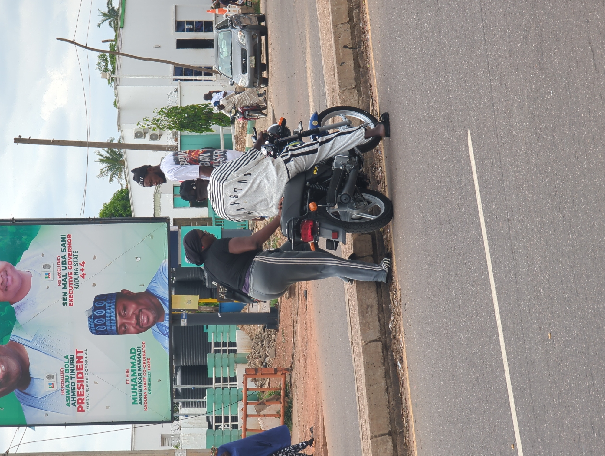The image depicts a street scene in Dadi Riba, Nigeria, under an overcast sky. A large political billboard occupies the upper left, featuring three male figures. From left to right, the billboard displays "ASIWAJU BOLA AHMED TINUBU PRESIDENT FEDERAL REPUBLIC OF NIGERIA," "SEN MAL UBA SANI EXECUTIVE GOVERNOR KADUNA STATE 4+4," and "RT. HON. MUHAMMAD ABUBAKAR MAMADI KADUNA STATE CO-ORDINATOR RENEWED HOPE."
In the foreground on the right, a person wearing a white and black striped long-sleeve shirt with "RAPSTAR" text and matching striped pants is seated on a blue and black motorcycle. Next to the motorcycle, a person in a black short-sleeve top, grey sweatpants with white side stripes, and black sandals is standing, facing the seated individual. A third person, wearing a white graphic t-shirt and a black beanie, is partially visible behind the motorcycle driver, looking towards the left.
The road is asphalt with a single white dividing line. A concrete curb separates the road from a dirt and gravel shoulder, where some debris is visible. On the far left, a person in a dark blue garment is walking away from the camera. Orange construction barriers and several black water storage tanks are visible behind the walking individual.
In the mid-ground right, a silver Toyota Matrix with a visible "KADUNA" license plate is on the road, alongside a red motorcycle. A person in a light-colored traditional garment is standing near the red motorcycle. White, modern-style buildings with blue-tinted windows and external air conditioning units are in the background. Palm trees and utility poles are also present.