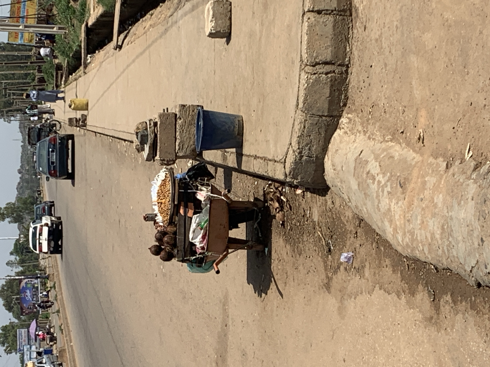 The image depicts a street scene in Jos, Nigeria. In the foreground, a wooden handcart laden with goods sits on the side of the road, adjacent to a concrete curb. The cart contains what appears to be a basket of nuts and several coconuts, along with various items wrapped in cloth. Behind the cart, the road stretches into the distance, occupied by several vehicles and pedestrians. The road is bordered by vegetation and buildings. The sky is overcast, suggesting midday or afternoon. The overall scene captures a typical street vending environment in Jos.