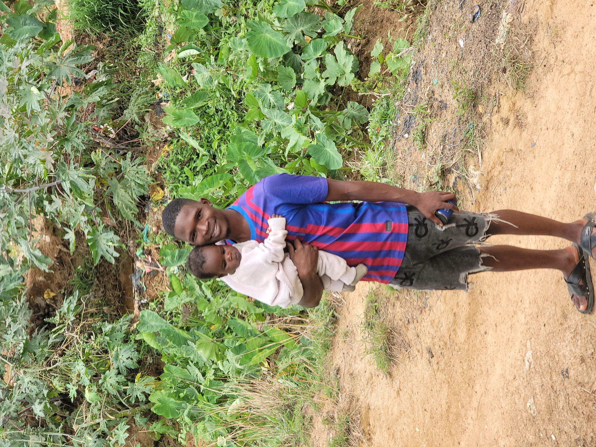 An adult male is holding a baby in front of foliage. The man is dark-skinned and wearing a blue and red striped t-shirt, dark shorts, and black sandals. He is holding the baby close to his chest with both arms. The baby is wearing white clothes. They are standing in a yard with mixed dirt and sand. There is a heavy presence of green foliage nearby. The scene is located near Barakin Akawo, Nigeria.