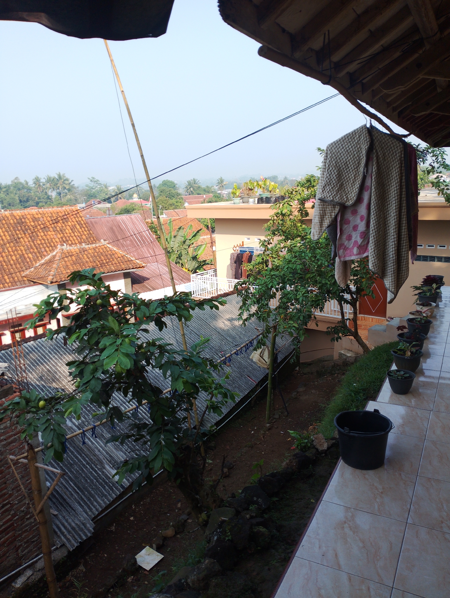 The image depicts an outdoor scene in Sukamanah, Indonesia. In the foreground, a tiled balcony extends into the frame, with several potted plants lining its edge. A black bucket sits on the tiles near the balcony's end. Beyond the balcony, a leafy tree with green fruit is prominently featured. The background reveals a residential area with orange-tiled roofs, some buildings, and lush green vegetation including palm trees. A clothesline with various garments hangs near a building, and a bamboo pole extends upwards into the sky. The sky is clear and pale blue, suggesting daytime. The overall impression is of a residential area in a tropical or subtropical climate.