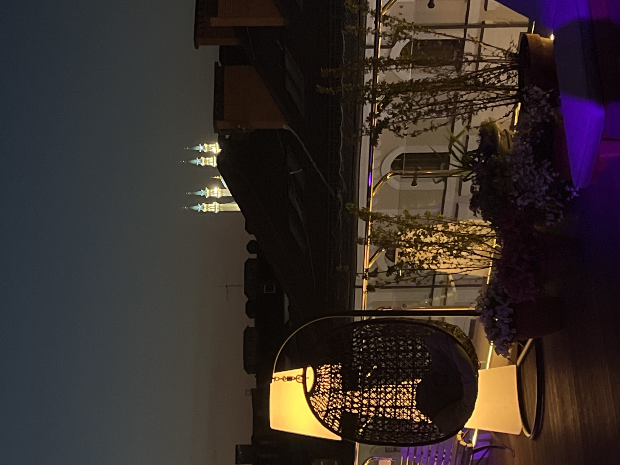 At dusk, a low-angle view from a rooftop in Kazan, Russia, focuses on a patio arrangement, including a hanging basket chair illuminated by a warm light source. The patio is adorned with plant life. In the distance, beyond the rooftop structures, the illuminated minarets of the Qolşärif Mosque are visible. The sky transitions from dark grey at the top to lighter near the horizon.