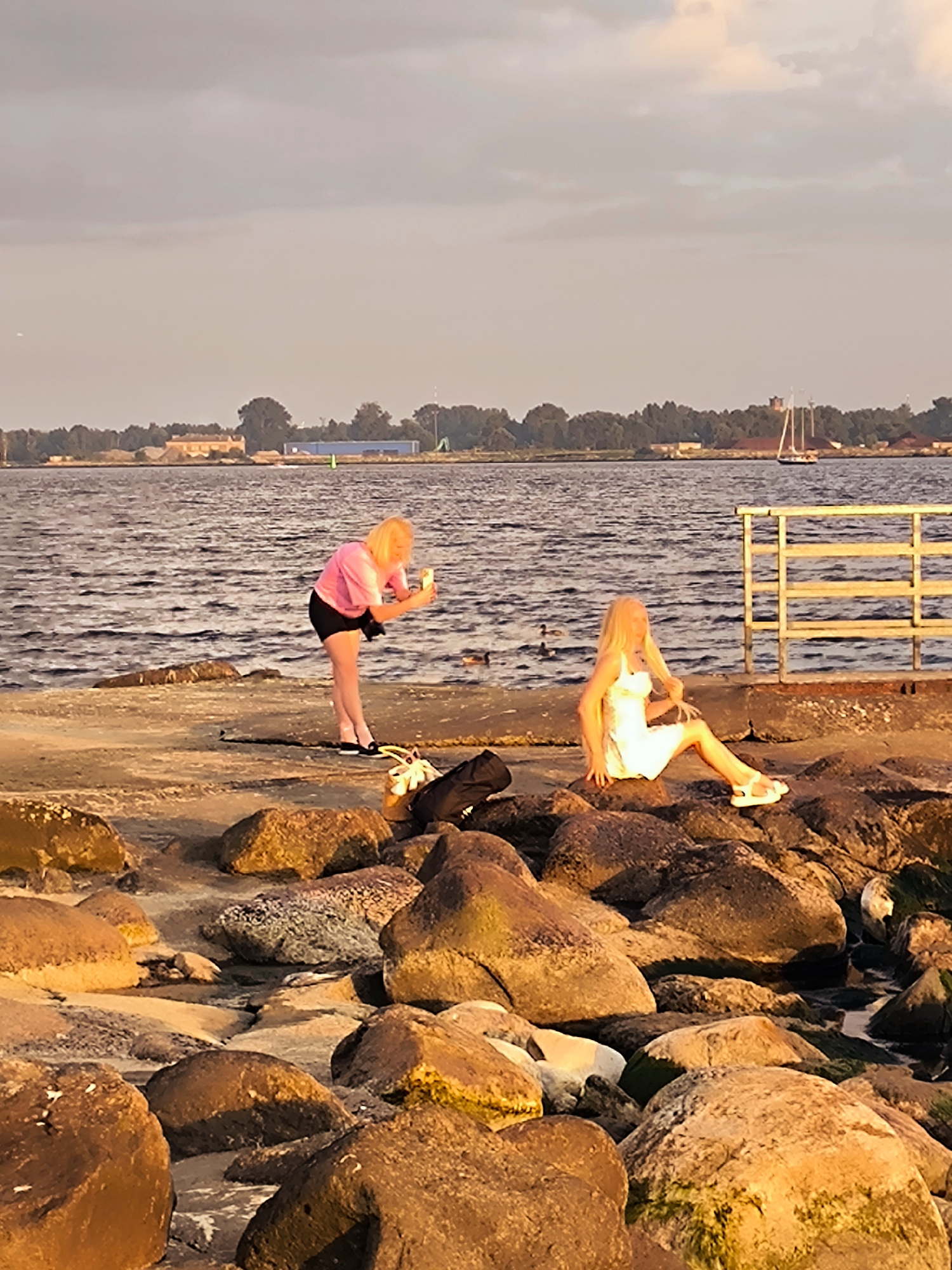 Two women are on a rocky shoreline by the water. One woman, with blonde hair and wearing a pink t-shirt and black shorts, is standing and holding a smartphone as if taking a picture. The other woman, also with blonde hair and wearing a white dress and sandals, is sitting on a large rock, looking to her left. The water is calm with gentle ripples. In the background, there is a line of trees and buildings along the horizon, and a sailboat is visible on the water. A yellow metal railing is present on the right side of the frame. The scene appears to be illuminated by the warm light of a sunset or sunrise. The location is identified as being near the Daugava River in Riga, Latvia.