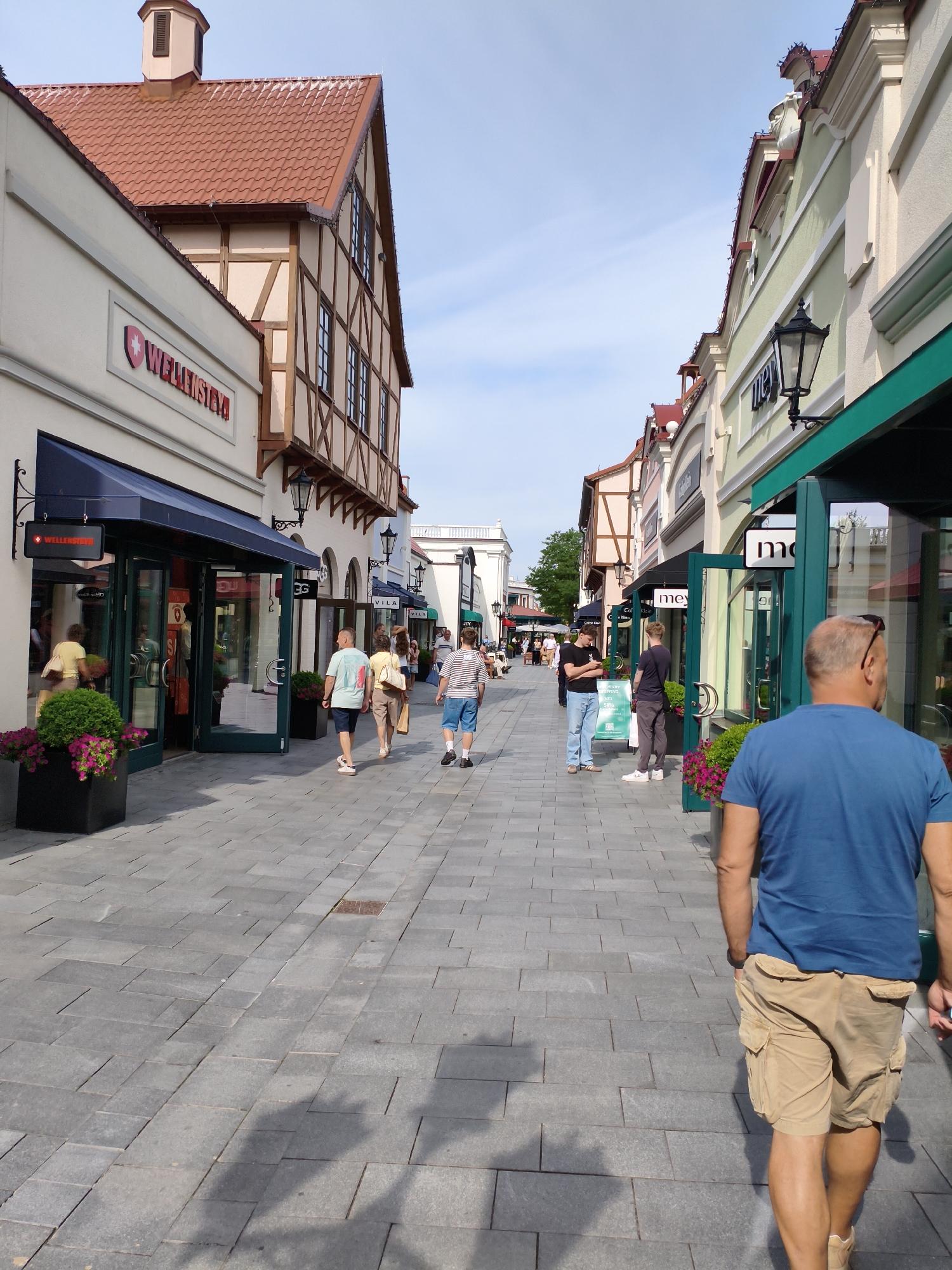 This is a photograph taken at the Designer Outlet Neumünster in Germany. The scene depicts a pedestrian walkway lined with shops. In the foreground, a man wearing a blue t-shirt and tan cargo shorts is walking away from the camera. Further down the walkway, other individuals are seen browsing shops or walking. The architecture of the buildings features a mix of styles, with some exhibiting half-timbered elements and others having a more modern facade with large display windows. The ground is paved with grey stone tiles. The sky is clear and blue, suggesting it is daytime and fair weather. Planters with pink flowers are visible near some of the shop entrances. The overall impression is that of a shopping district.