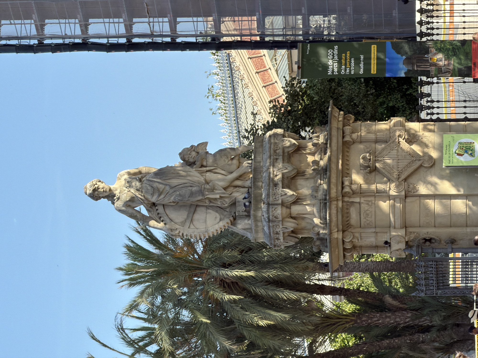 A sunlit outdoor scene in Barcelona, Spain, features a monumental stone statue atop a multi-tiered pedestal. The statue depicts a female figure, possibly allegorical, with drapery and a circular object with spokes. A small winged cherub is positioned beside her. The pedestal is adorned with classical architectural elements and decorative carvings. In the background, palm trees and lush greenery are visible, alongside a building with a distinctive glass roof and ornate trim, likely a greenhouse or conservatory. A tall, modern structure covered in scaffolding is also present on the right side of the frame. A vertical banner on the right provides information about "Més de 600 parcs i jardins" and includes a website address. Several pigeons are perched on the pedestal. The sky is clear and blue. A few people are visible in the lower portion of the image, near a decorative iron fence.
