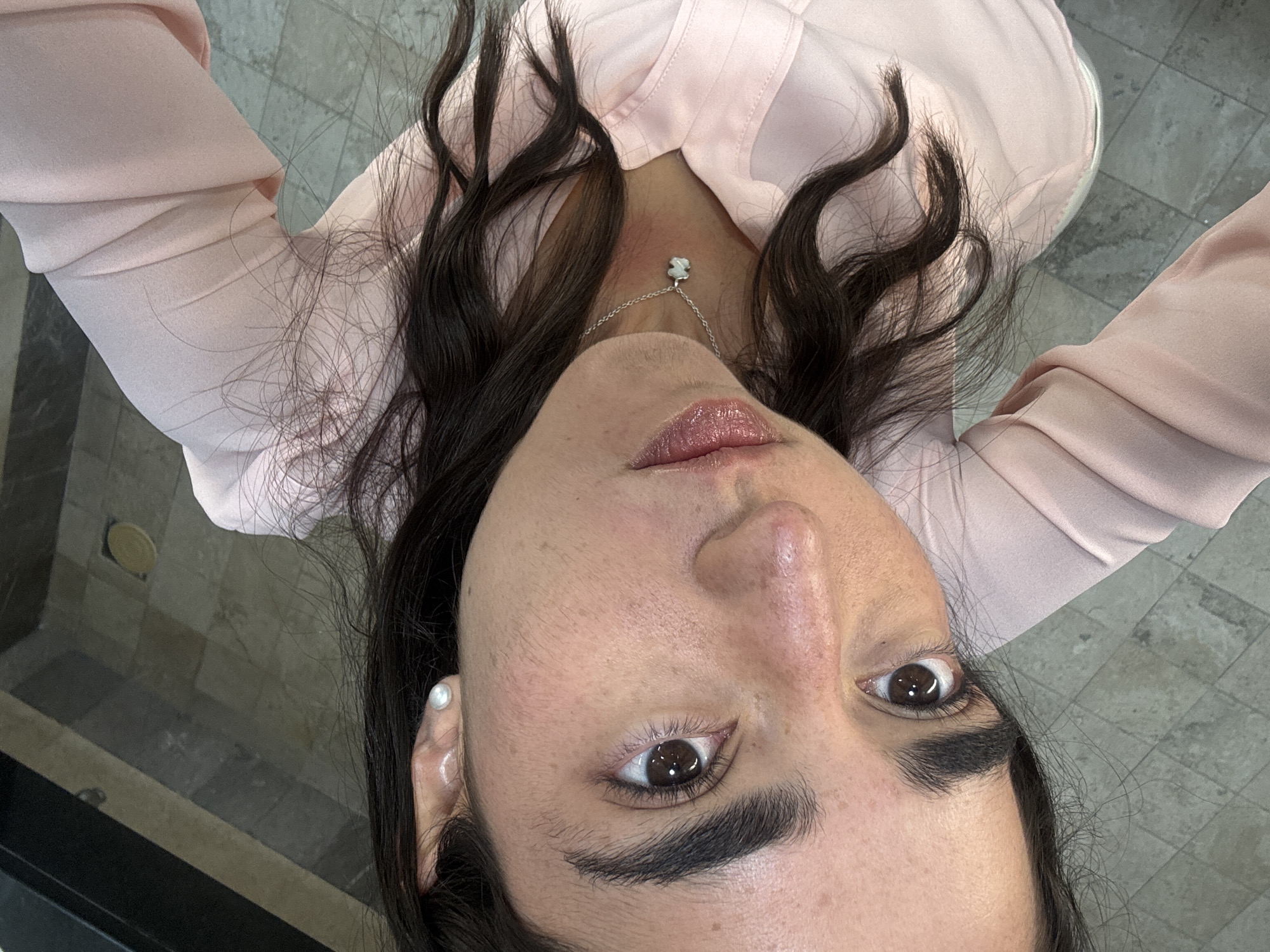This close-up, high-angle selfie captures a person looking directly upwards into the camera. The subject is a young adult with a medium-dark skin tone, dark, wavy hair that frames their face, and striking dark brown eyes. They are wearing a light pink, collared button-up shirt. A delicate silver-colored necklace with a small, white, bear-shaped charm, reminiscent of the Tous brand logo, rests on their neck. A pearl stud earring is visible in their right ear. Their lips are slightly parted and have a glossy finish. The person's expression is neutral and direct.
The background reveals an indoor setting with a tiled floor, featuring a pattern of light grey and darker grey/black square tiles. The floor appears polished, with visible reflections in several areas. This setting suggests a contemporary interior, possibly within a building or commercial space in Mexico City, Mexico. No discernible text is visible in the image.