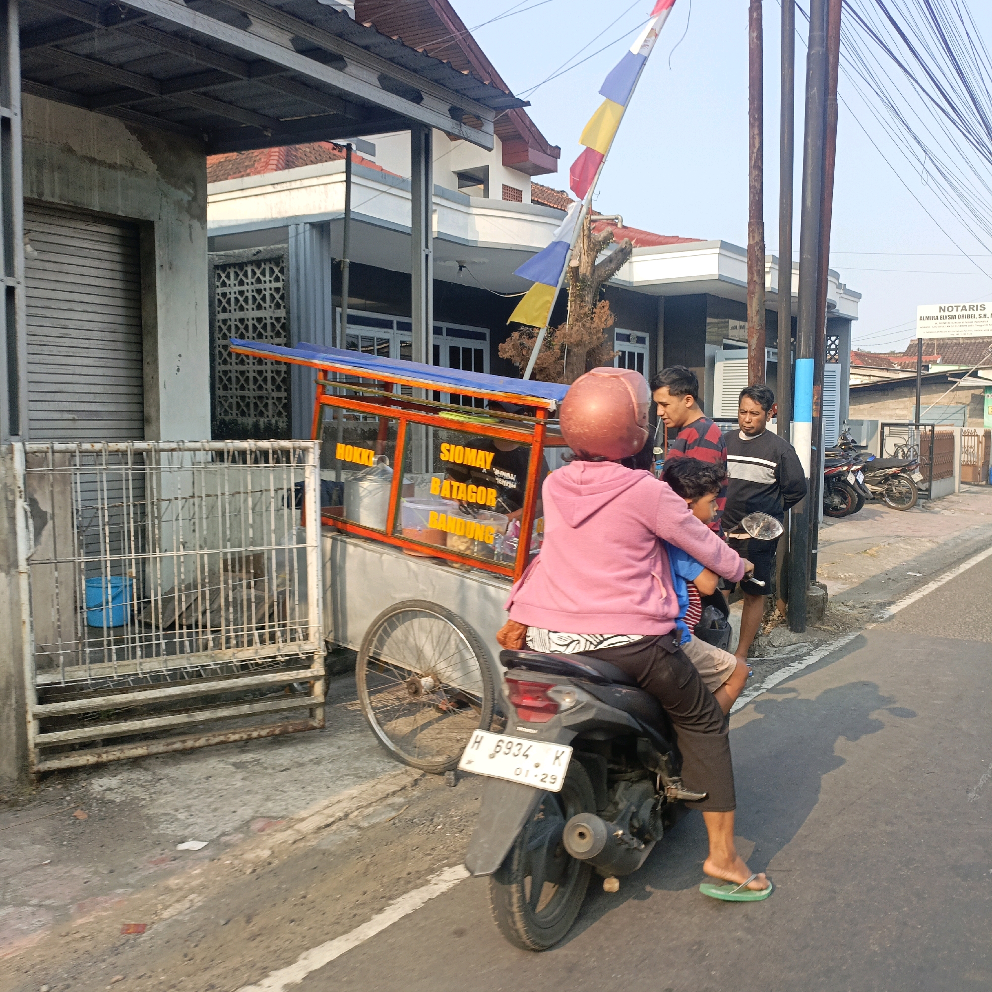 This is a medium-shot, eye-level photograph taken outdoors during the day in Salatiga, Indonesia, capturing a street scene with a food cart and people interacting.
In the foreground, a person wearing a pink hoodie and a red helmet is seated on a black motorcycle, facing away from the camera. They are wearing black pants and green flip-flops. A child in a blue shirt is seated in front of them on the motorcycle. The motorcycle's license plate is visible, showing "H 6934 K" and "01-29".
To the left of the motorcycle is a food cart with an orange frame and a blue tarp. The cart has glass panels displaying text that appears to read "HOKKI", "SIOMAY", "BATAGOR", and "BANDUNG". Inside the cart, there are metal containers and plastic tubs. A metal fence with a blue bucket behind it is situated to the left of the food cart.
Behind the food cart, three men are standing on the sidewalk. The man on the far right is wearing a black long-sleeved shirt with white stripes and black shorts. The man in the middle is wearing a striped shirt with red, black, and blue colors, and black shorts. The man on the far left, partially obscured, appears to be wearing a black and white striped shirt.
Behind the people and the food cart, there are buildings with tiled roofs. A national flag with red, white, yellow, and blue sections is visible on a pole. Various utility poles and a tangle of electrical wires are prominent in the background, along with a white sign that reads "NOTARIS ALMIRA ELYSIA ORIBEL, S.H., M.Kn." and other text. Several motorcycles are parked on the side of the road in the distance. The ground surface is asphalt and concrete. The lighting suggests a bright, possibly sunny day, with shadows cast on the road.