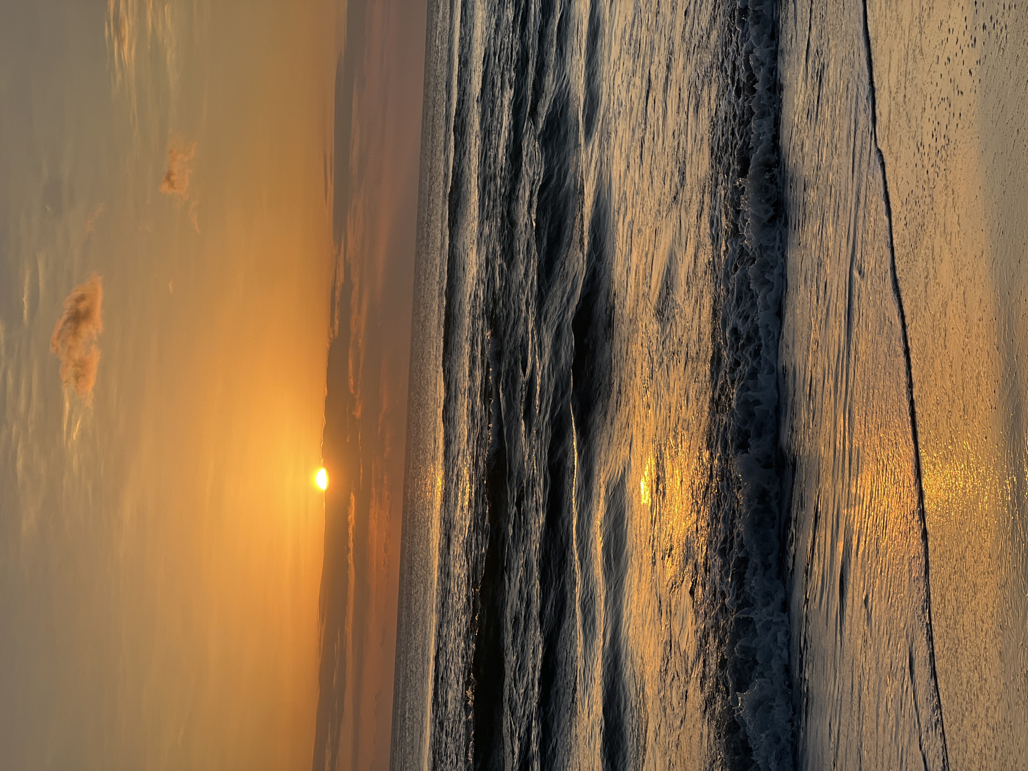 The image depicts a serene sunset over the ocean, casting a warm golden glow on the water and sky. The sun, a bright orb, is partially obscured by a band of clouds near the horizon, creating a dramatic effect. Gentle waves roll towards the shore, reflecting the vibrant colors of the sunset. The beach in the foreground appears wet, with the receding tide leaving a shimmering surface. The overall atmosphere is peaceful and tranquil. This scene is likely located on the coast of Kuta, Indonesia, a popular tourist destination known for its beautiful beaches and stunning sunsets.