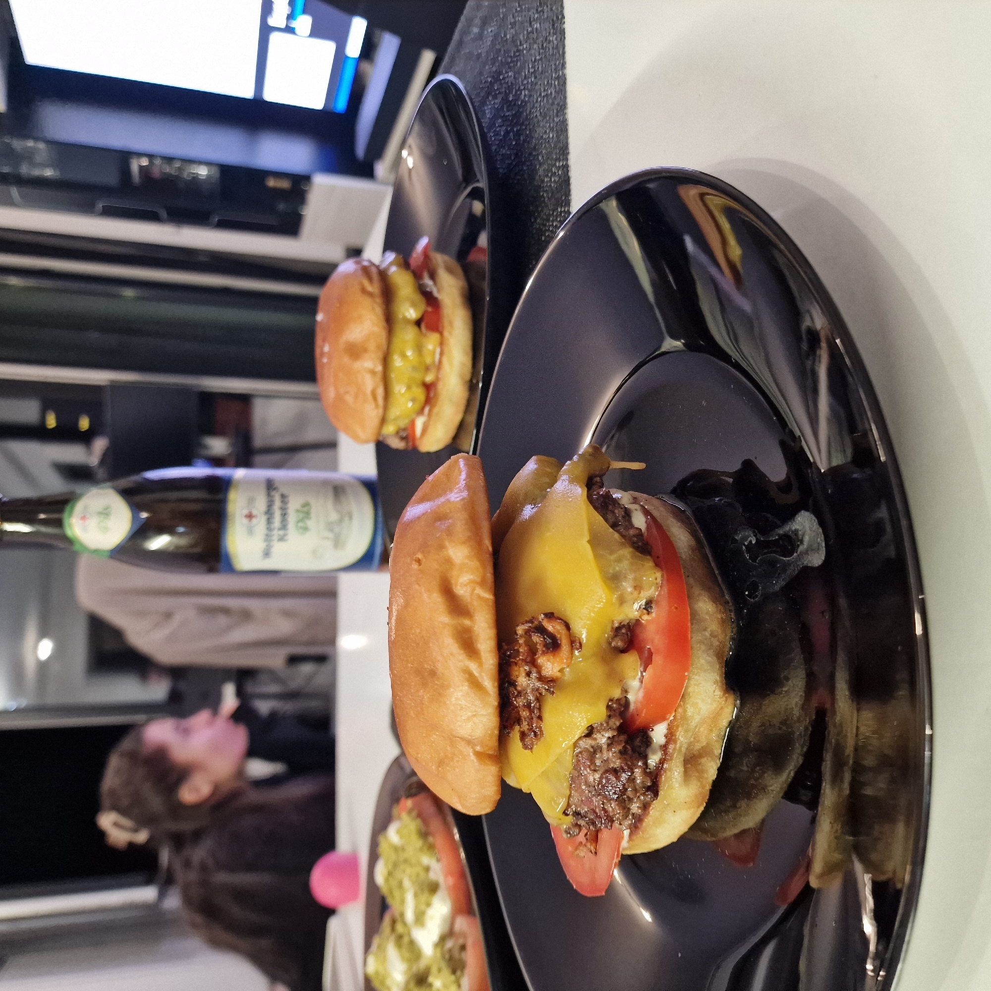 The image shows two open-faced cheeseburgers on black plates. A bottle of "Weltenburger Kloster Pils" beer is visible in the background, along with a person sitting at the table. The location is likely in Sofia, Bulgaria based on the beer. The burgers appear to contain beef patties, cheese, and tomato slices. Some sort of sauce or condiments is also visible. The background suggests an indoor setting, possibly a restaurant or dining area.