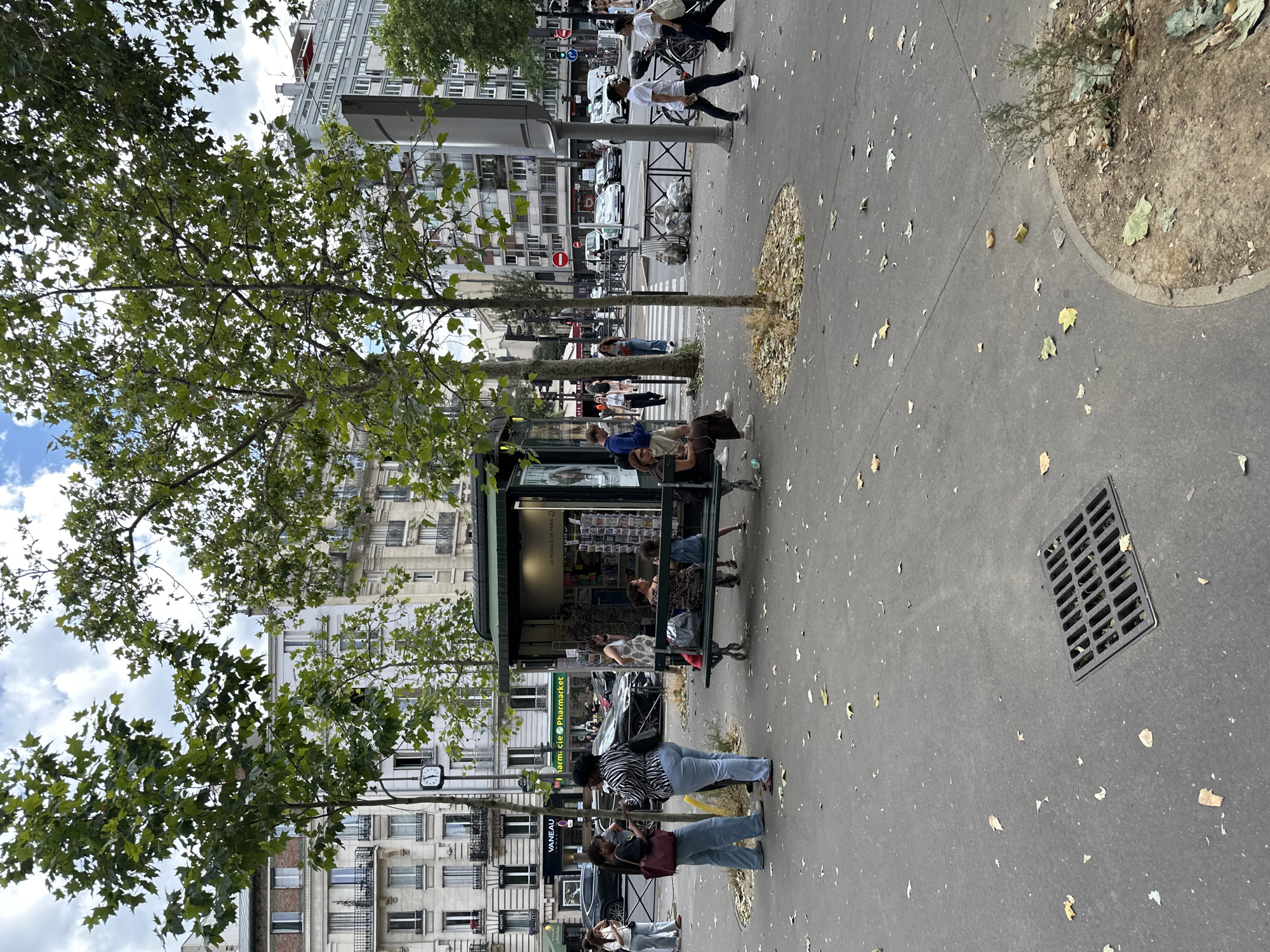 The image depicts a street scene in Paris, France. The perspective is from ground level, looking across a sidewalk and a street with a kiosk, buildings, and trees. The time of day appears to be daytime, with a cloudy sky.
On the sidewalk, several people are present. Some are seated on a bench near a kiosk that appears to be selling newspapers and magazines. Others are standing or walking. One woman in a black and white striped shirt and blue jeans is looking at her phone, while another woman next to her, in a red top and jeans, also appears to be looking at her phone. Further down the sidewalk, a group of people are near a street crossing, with one person in a white t-shirt and black pants walking away from the camera. A man in a white t-shirt and black pants is visible in motion, suggesting an activity like dancing or a performance. Cars are visible on the street, indicating an urban environment.
Notable details include a metal grate in the pavement and scattered fallen leaves on the ground, suggesting it might be late summer or early autumn. The buildings lining the street are multi-story, with balconies and awnings, characteristic of Parisian architecture. Some text is visible on signs, including "VANEAU" and "Pharmacie," indicating commercial establishments. The overall atmosphere seems casual and typical of a bustling city street.