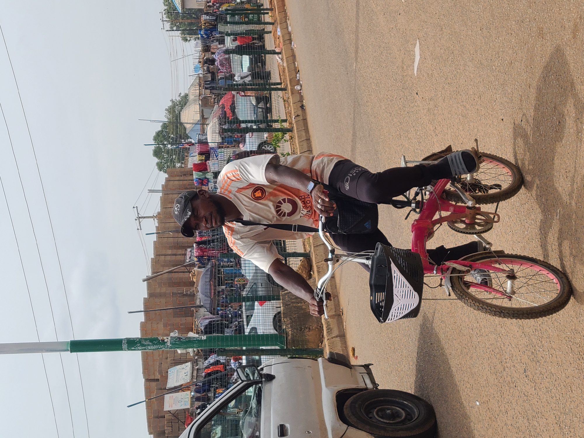 A man is riding a pink bicycle on a paved road in an urban environment. He is wearing a black cap, a white and orange shirt, black leggings, and a watch. He has a black bag strapped across his body. The bicycle has a black basket attached to the front. A white van is parked to the left of the road. In the background, there is a row of stalls or shops selling clothing and other items. There are utility poles with power lines and a green and white street sign pole on the left side of the frame. The sky appears overcast. The setting seems to be a busy street or market area.