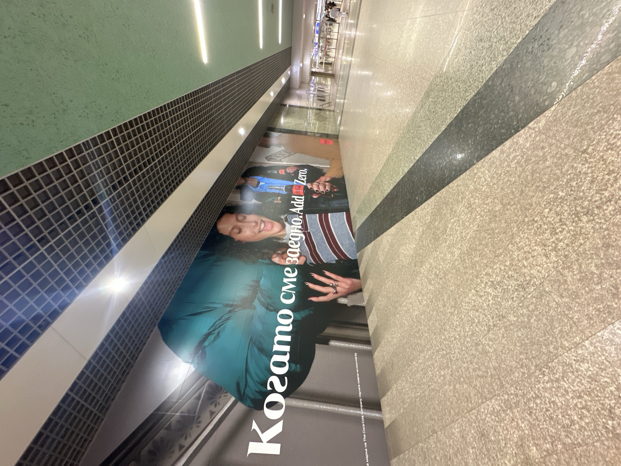 Here is a detailed description of the image:
**Scene:** The image shows a wide, brightly lit indoor space that appears to be a corridor or walkway, likely within an airport or large commercial building. Along the left side, there's a wall with a large advertisement. The floor is tiled with a light speckled pattern, featuring a diagonal dark gray accent stripe.
**Advertisement:** The ad on the wall features a woman with the text "Когато сме заедно. Add Zero." ("When we are together. Add Zero.") in Bulgarian and English. Additionally, there is smaller text at the bottom that indicates "Coca-Cola Company".
**Details:** The ceiling has long, recessed fluorescent lights running parallel to the corridor's length. In the distance, there are people visible, further suggesting a public area.
**Location:** Given the language in the advertisement and the general architectural style, it's plausible that this scene is located in Sofia, Bulgaria. Airports and large commercial centers in Sofia often feature advertisements in Bulgarian and English.