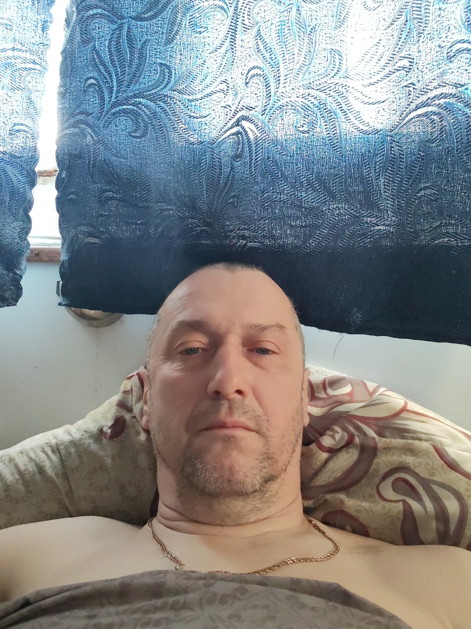 The image shows a middle-aged man lying down on patterned bedding. He has a shaved head, stubble on his face, and is wearing a gold chain around his neck. He appears to be under a gray blanket. Behind him are blue patterned curtains partially covering a window. The location is Unknown, Unknown.
