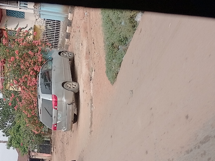The scene depicts a static outdoor environment in Orie Uratta, Nigeria, under clear daytime lighting. A silver Toyota Sienna minivan, with visible license plate characters "BAY 782", is parked on unpaved light brown earth, angled towards the right. The vehicle's rear bumper shows damage, with a missing section on the lower left. Behind the minivan, a prominent bush with numerous pink flowers (consistent with bougainvillea) partially covers a light yellow building. A white metal fence, topped with barbed wire, is situated in front of the building. To the immediate right of the minivan, square concrete blocks line what appears to be a property boundary. Further right, a light blue building with a patterned blue window is visible. The foreground consists of an unpaved dirt path, with a patch of green ground cover visible on the right. No individuals or active events are depicted.