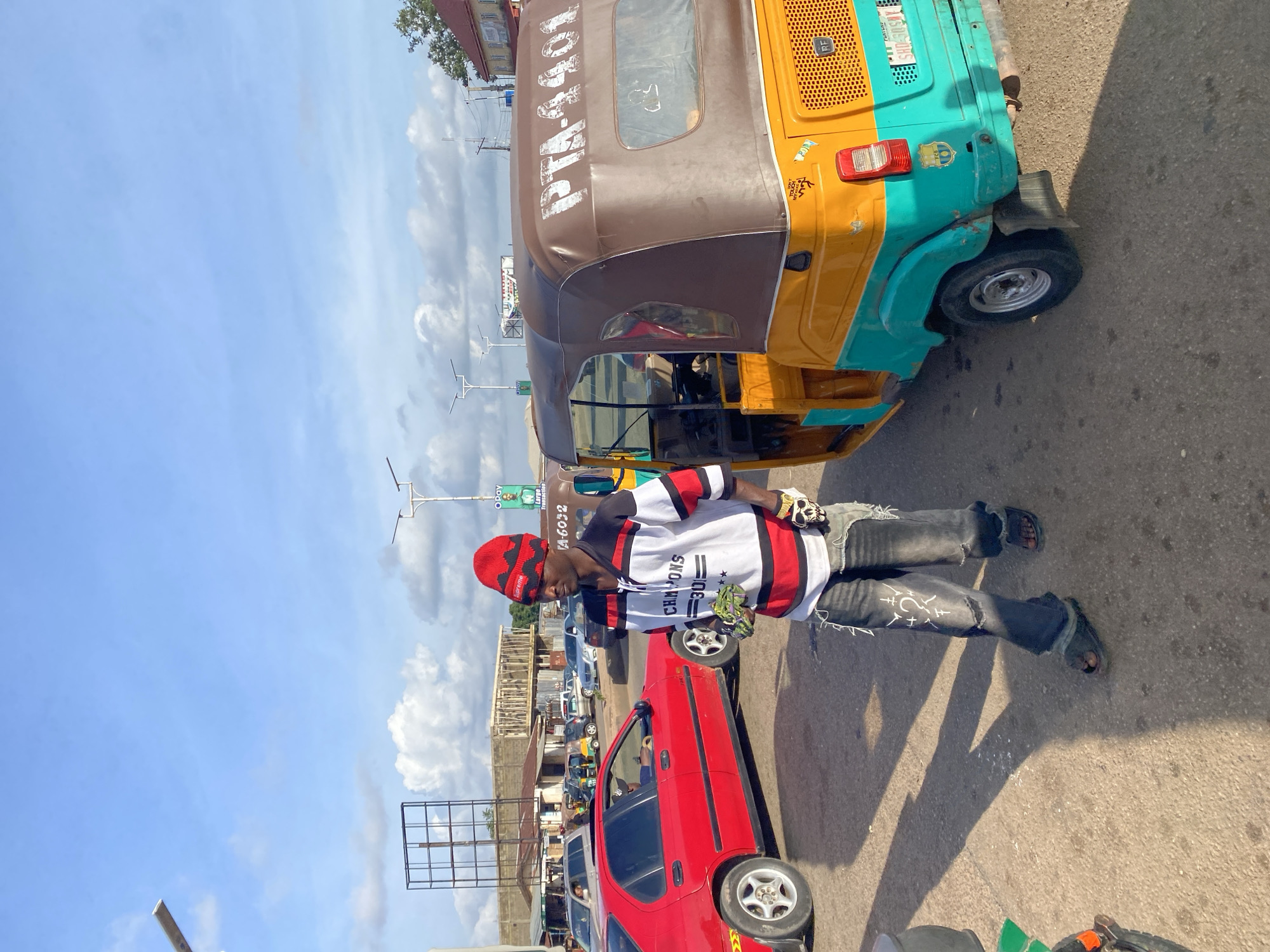 A man wearing a red and black patterned beanie, a white and black striped shirt, and ripped black jeans is standing in a street in Gura Top, Nigeria. He is holding a small green object in his left hand and has a skull accessory on his right wrist. To his right, a yellow and turquoise auto-rickshaw with the license plate "PTA-4401" is parked. A red sedan is visible to his left. The scene appears to be a street with other vehicles and buildings in the background. The sky is blue with scattered clouds.
