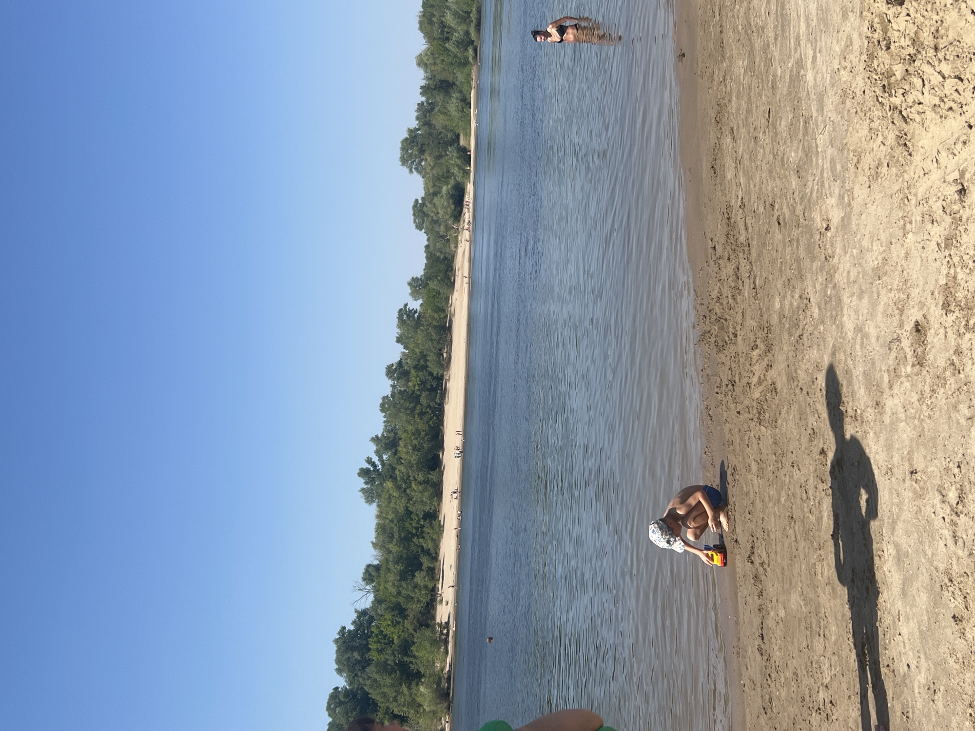 The image depicts a sunny day at the Kilyakovka River in Russia. A sandy beach lines the left bank of the wide, calm river, backed by a dense forest of green trees. The sky is a clear, bright blue, suggesting pleasant weather.
Several people are visible enjoying the river and beach. In the water, a woman in a black bikini is standing, looking towards the camera. Further down the river, a person is wading. Closer to the shore, a young boy wearing a patterned cap and blue shorts is crouching on the sand, playing with a colorful toy truck near the water's edge. His shadow stretches long across the sand, indicating it is likely late afternoon or early morning. Other beachgoers are scattered along the sandy bank in the distance. The overall scene conveys a relaxed and summery atmosphere, with people engaging in typical riverside activities like swimming and playing. There is no visible text in the image.