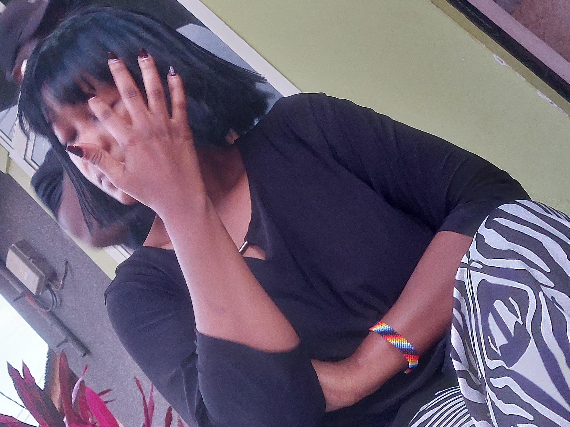A person is sitting outdoors in Dar es-Salaam, Tanzania. The individual, who appears to be a woman, has dark hair with bangs, dark nail polish, and a dark-colored shirt. She is partially obscuring her face with her hand. Her pants are patterned with black and white zebra-like stripes. She is wearing a colorful beaded bracelet. The background includes a light green wall, a section of a building with an electrical box, and some red plants.