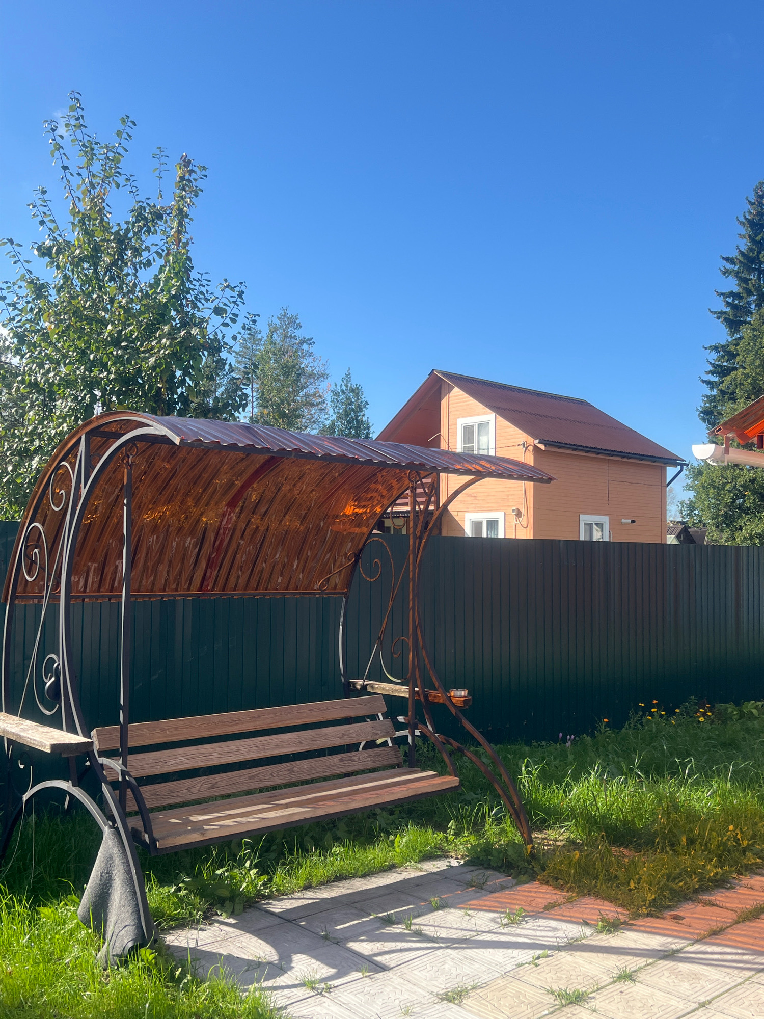 This is a medium shot, eye-level, outdoor photograph of a swing set in a grassy yard on a sunny day. The swing set is in the foreground, with its wooden seat visible. The swing set has a metal frame with decorative swirls. A brown, corrugated awning is attached to the top of the swing set. In the background, a dark green metal fence separates the yard from a residential area. Behind the fence, an orange wooden house with white windows and a dark brown roof is visible. The sky is clear blue. There are trees on either side of the house, and a portion of a large evergreen tree is visible on the far right of the image. The location is identified as Otradnoye, Russia.