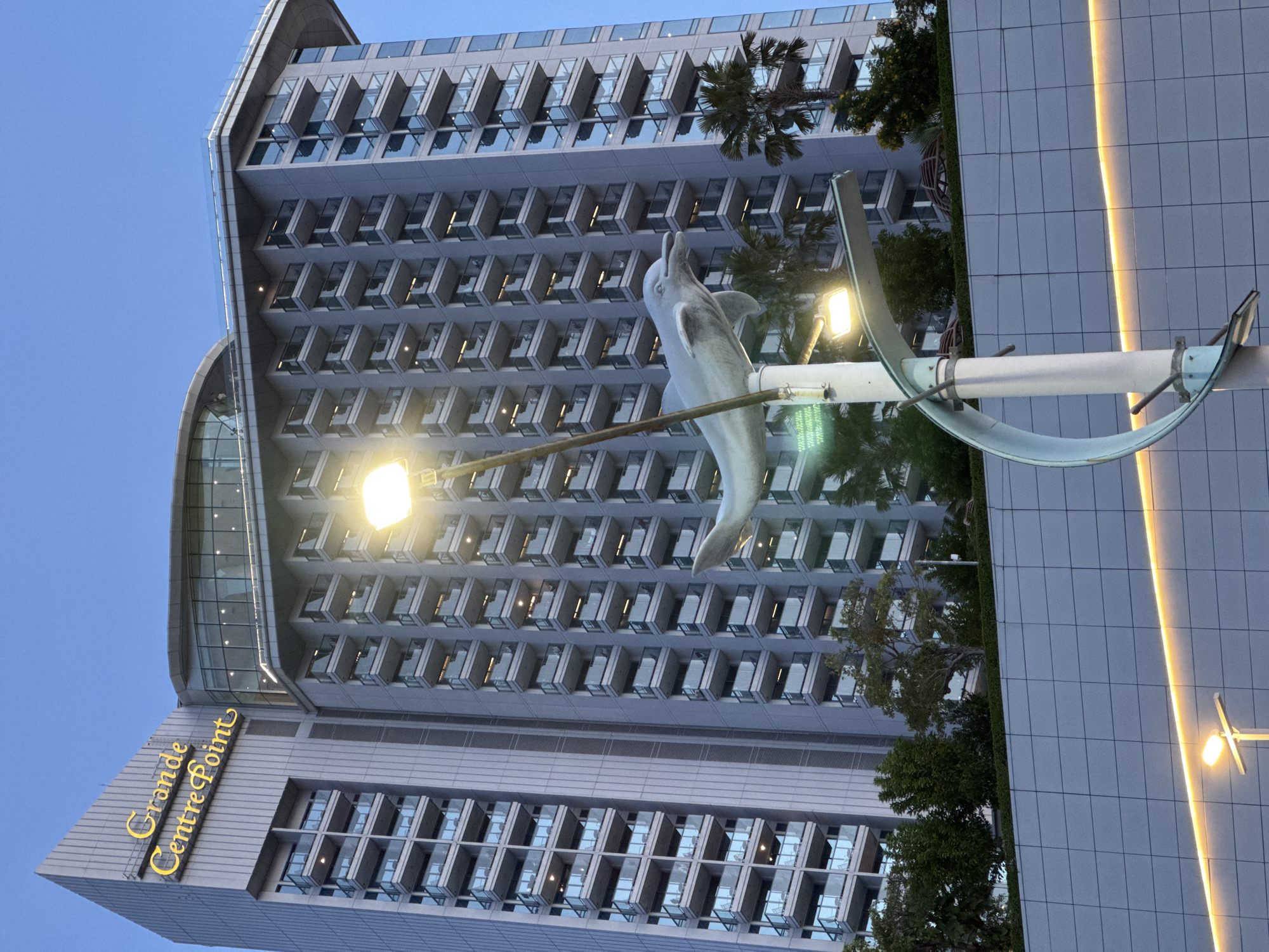 The image shows the Grande Centre Point Hotel Pattaya, a tall hotel building located in Pattaya City, Thailand. The hotel has a modern architectural design with many balconies and glass windows. In the foreground, a street lamp with a dolphin sculpture is visible. The sky is clear and blue, suggesting it is either early morning or late afternoon. The hotel is situated in a well-developed urban area, with other buildings and palm trees in the background. The lighting suggests the scene is during twilight.